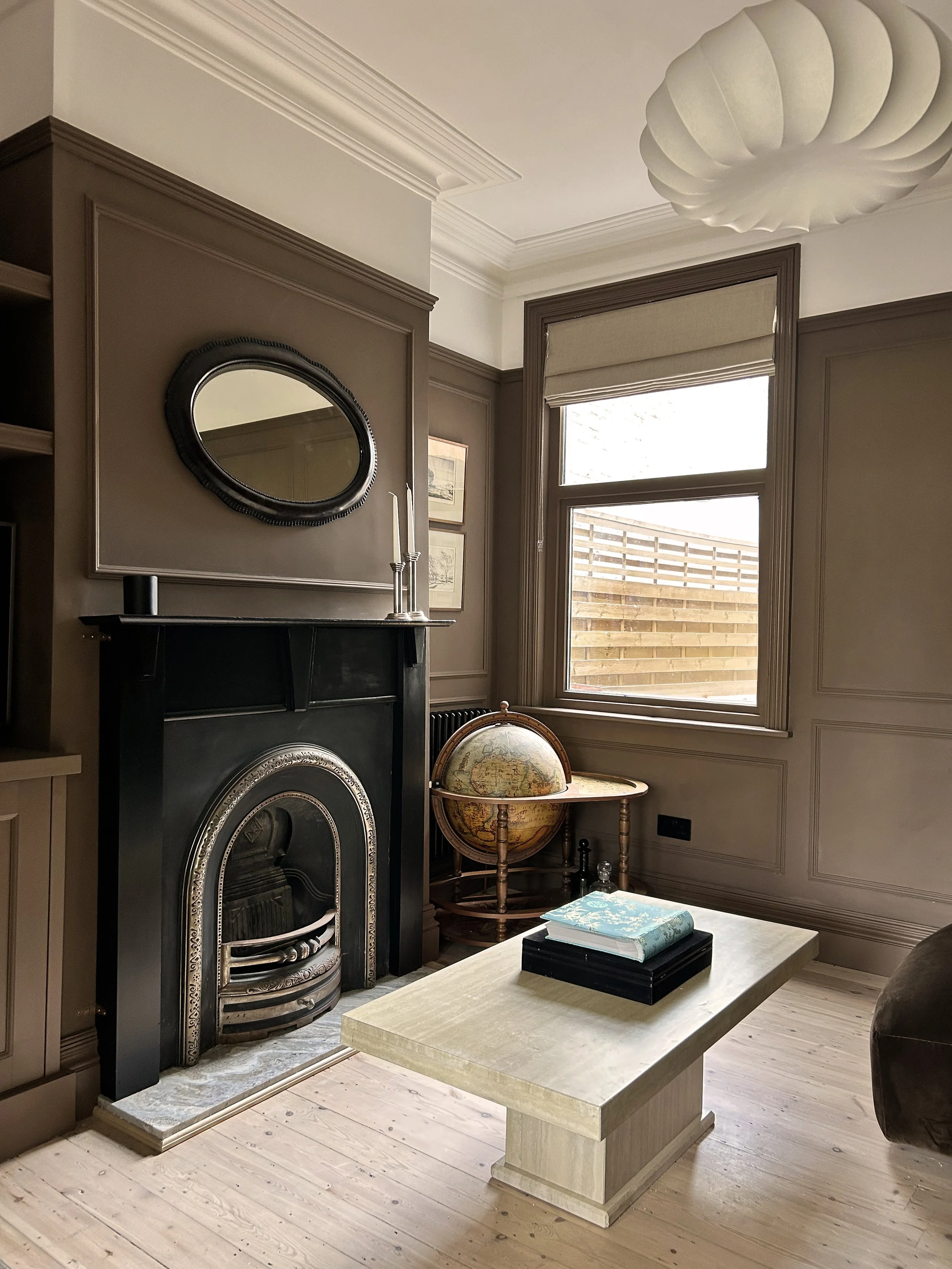 Interior of a moody, dark living room with a black fireplace, a globe on a small round table, a stack of books on a beige coffee table, a window with a roman blind, framed pictures on the wall, and a modern white ceiling fixture.