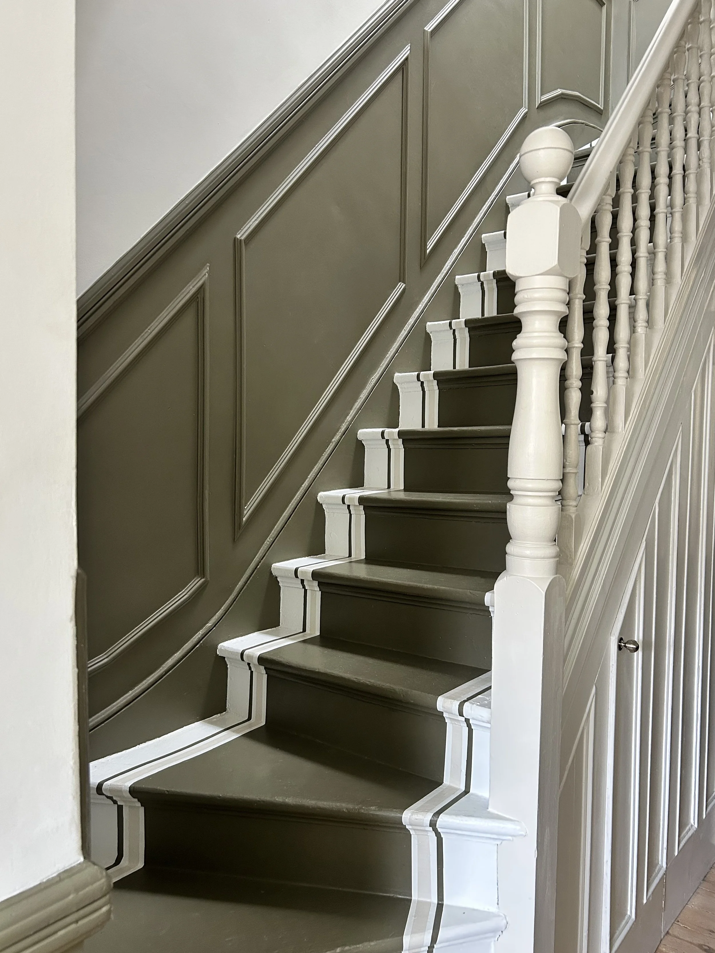 Interior of a staircase with dark-colored steps, white risers, and a white wooden railing with balusters, against a wall with decorative moldings painted in dark gray, with the wall above painted in light cream.