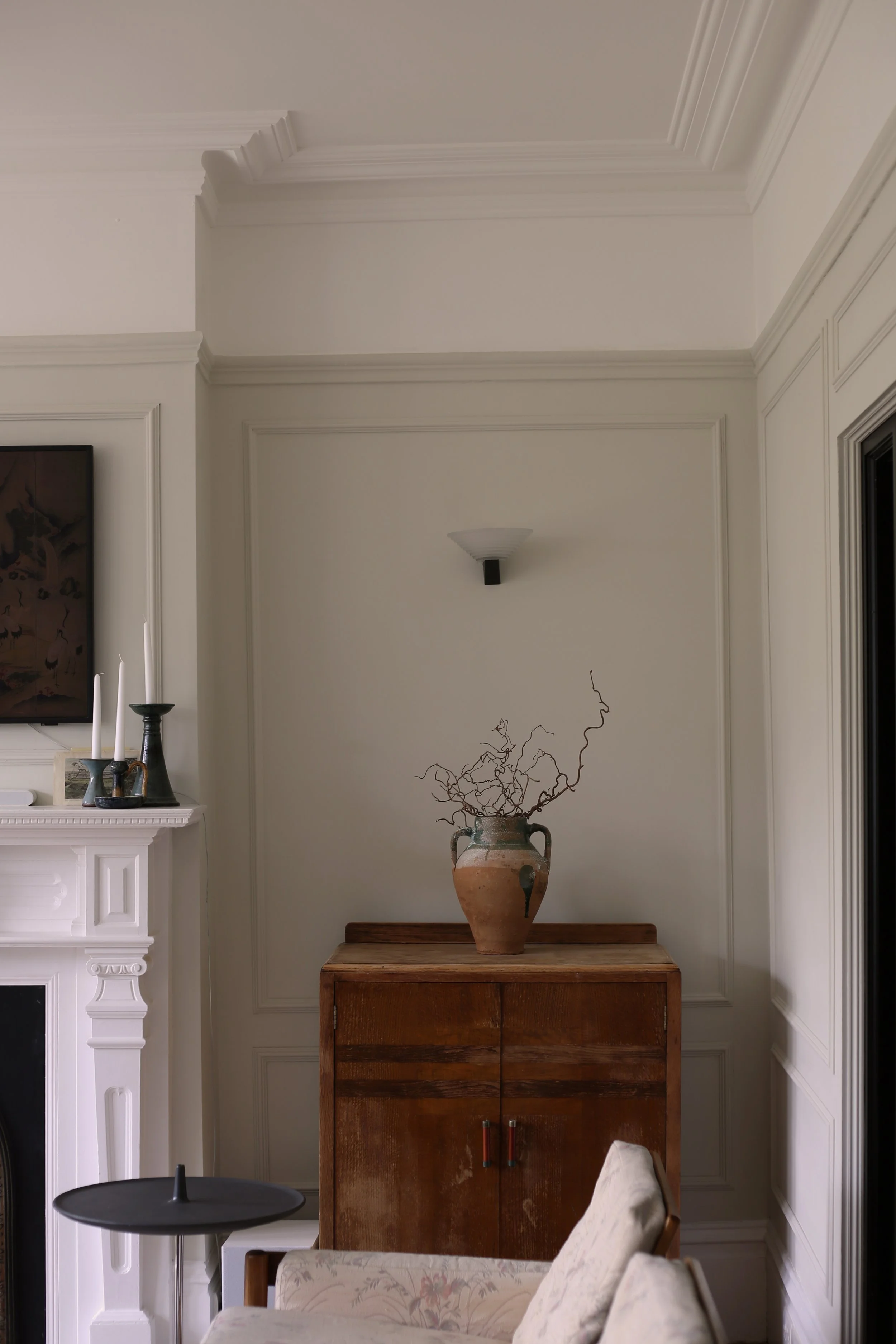 Living room corner with white walls, decorative molding, and a wooden cabinet with a large terracotta vase filled with twisted dried branches on top. A wall sconce is mounted above the cabinet, and part of a white fireplace with candles and a framed picture are visible on the left. A floral upholstered chair and a black side table with a sculpture are in the foreground.