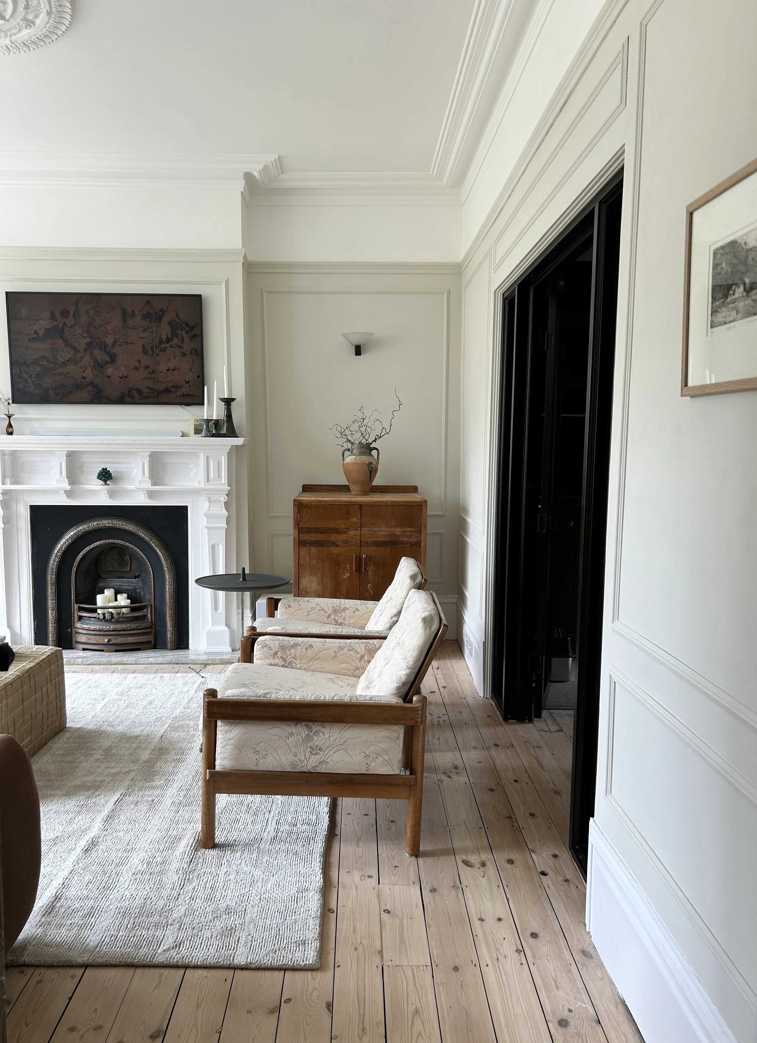 Living room with neutral walls, a fireplace with candles, wooden flooring, beige upholstered chairs, a small black side table, a wooden cabinet with a decorative vase, and framed artwork on the wall. Edwardian architectural details are all visible.