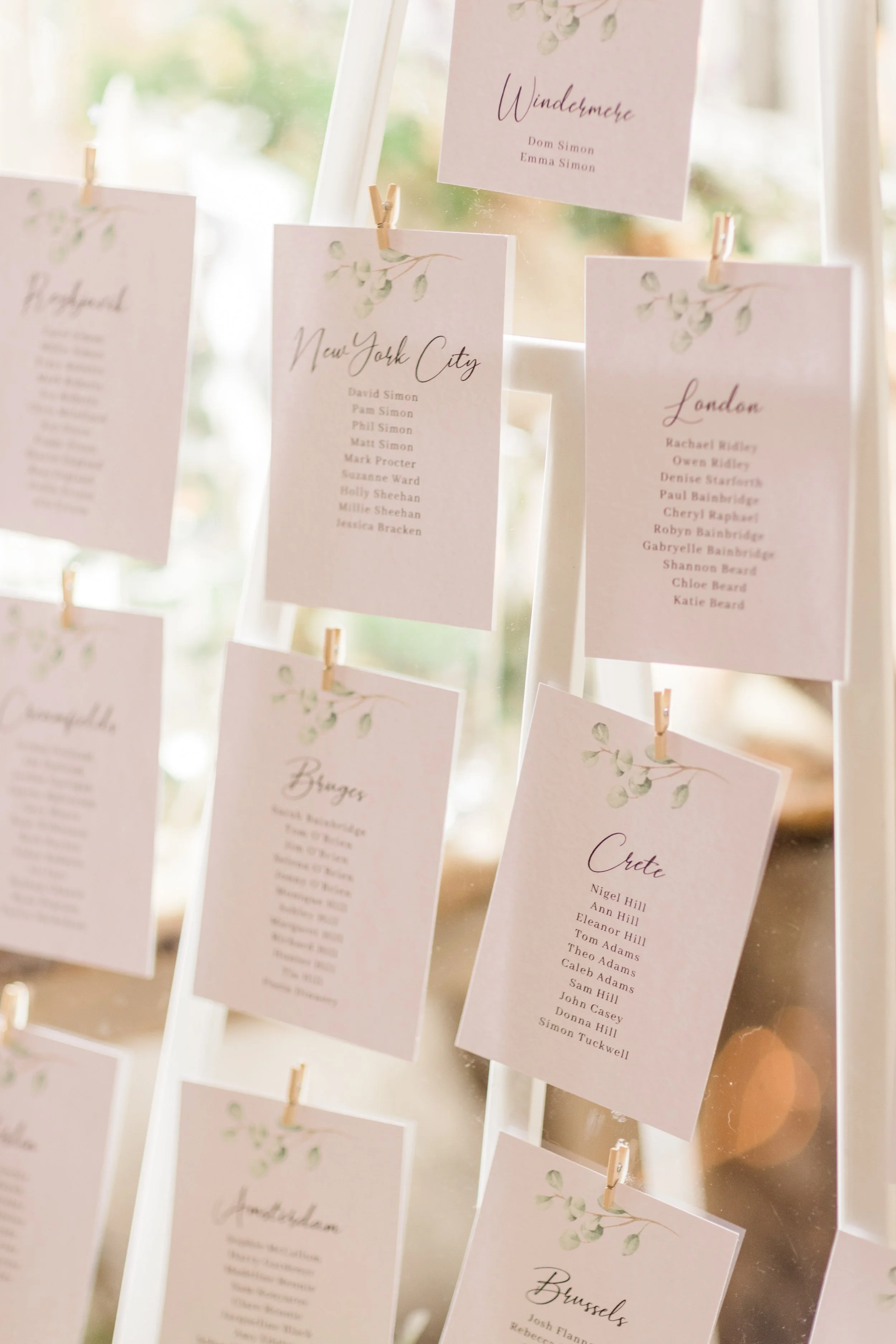 Wedding seating chart with cards displaying guest lists from various cities, attached with clothespins on an acrylic display stand.