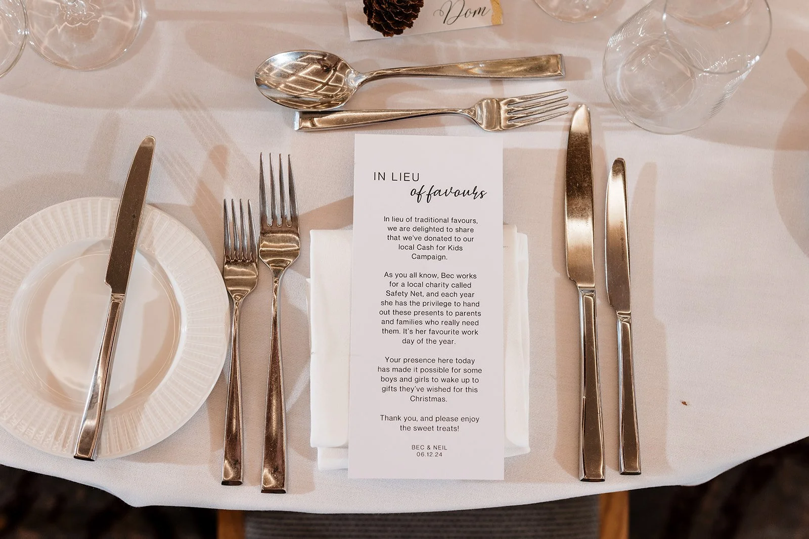 A formal table setting at a wedding or event, with silverware including a knife, fork, spoon, and a smaller fork, a white plate, a white napkin, a printed program or menu, and clear glasses, on a white tablecloth.
