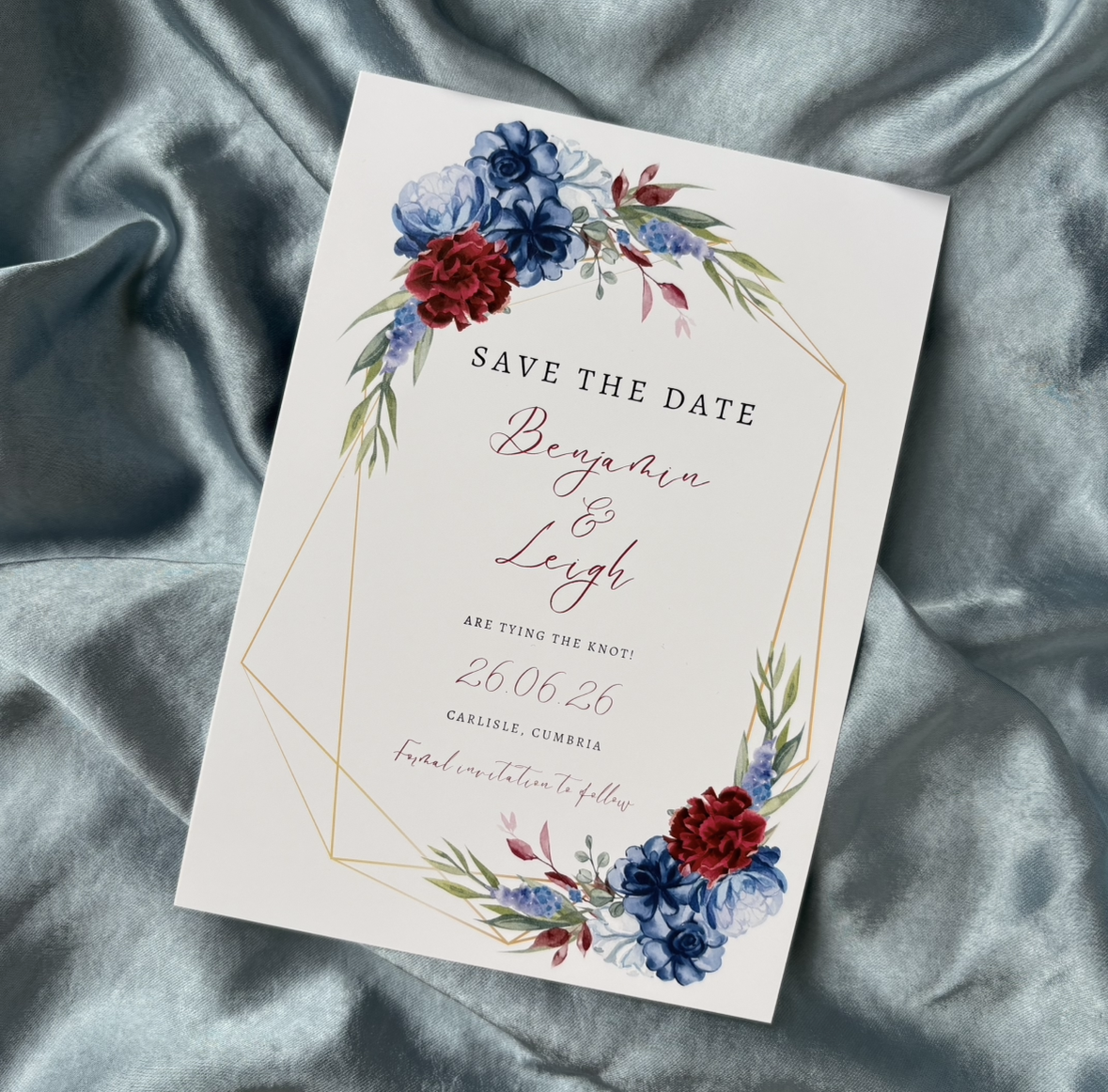 Wedding save the date card with floral designs in blue, burgundy and green, gold geometric border, and elegant text, placed on a light blue satin fabric background. Tithe Barn Carlisle Cumbria