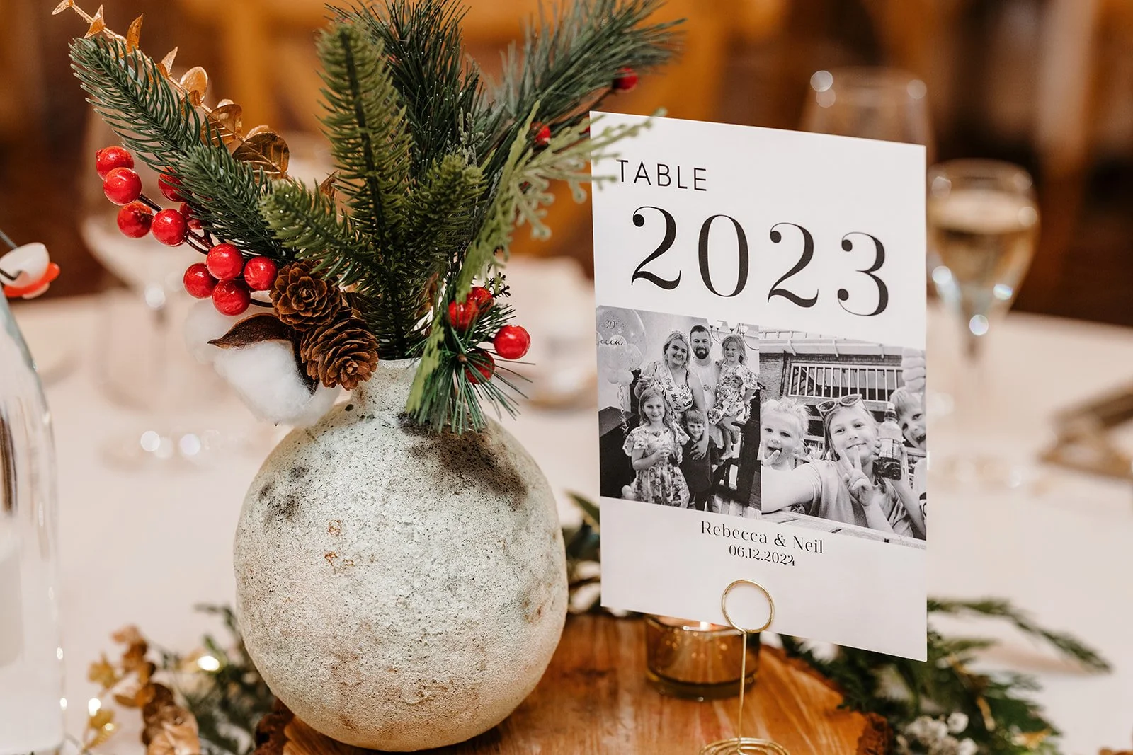 Table centerpiece with a small greenery arrangement in a textured round vase, alongside a personalised table number card labeled '2023' with black and white family photos, names 'Rebecca and Neil,' and the date '06.12.2023'.