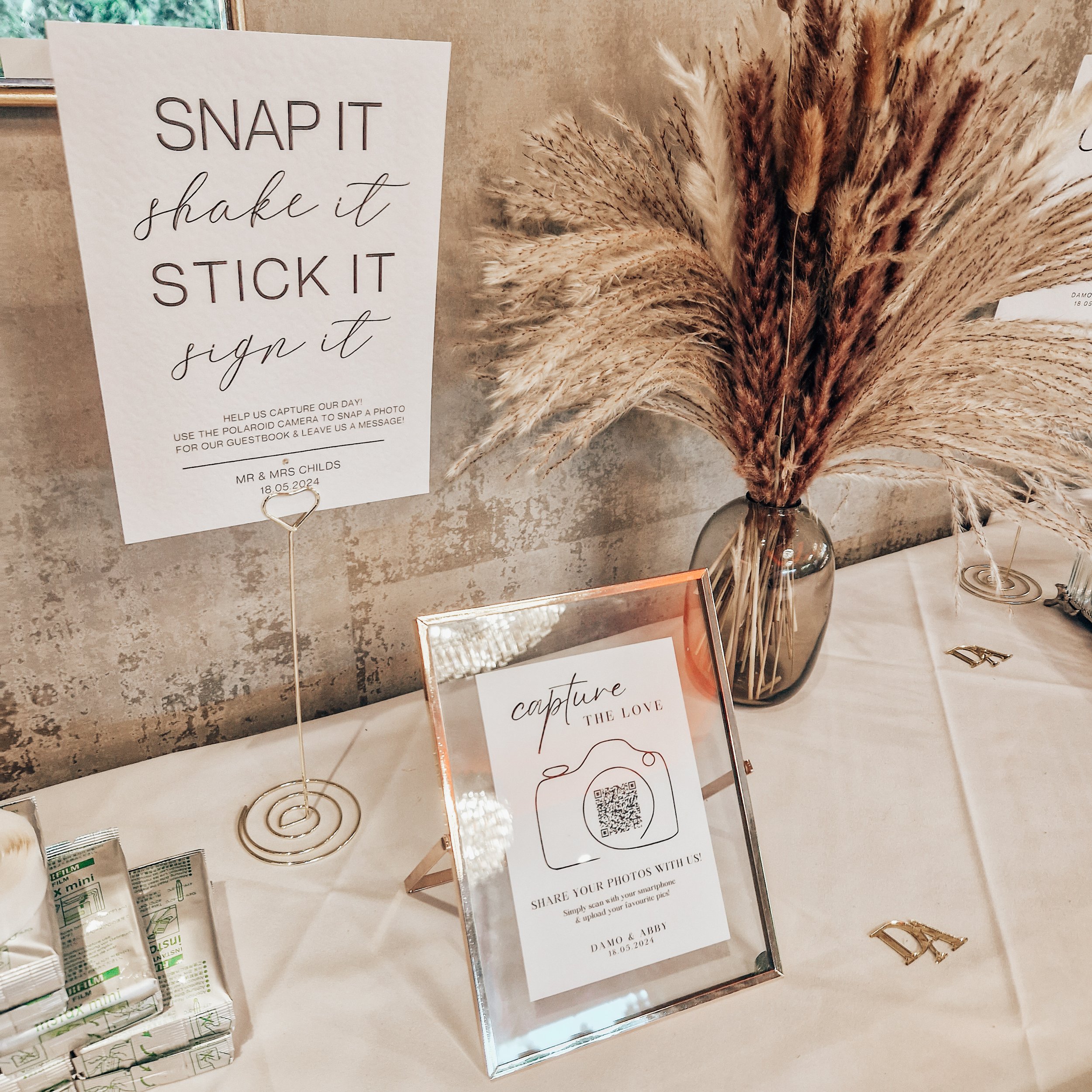 Table with a sign instructing to snap, shake, stick, and sign, a frame with a camera illustration and a QR code for sharing photos, a vase with dried pampas grass, and small decorative items.