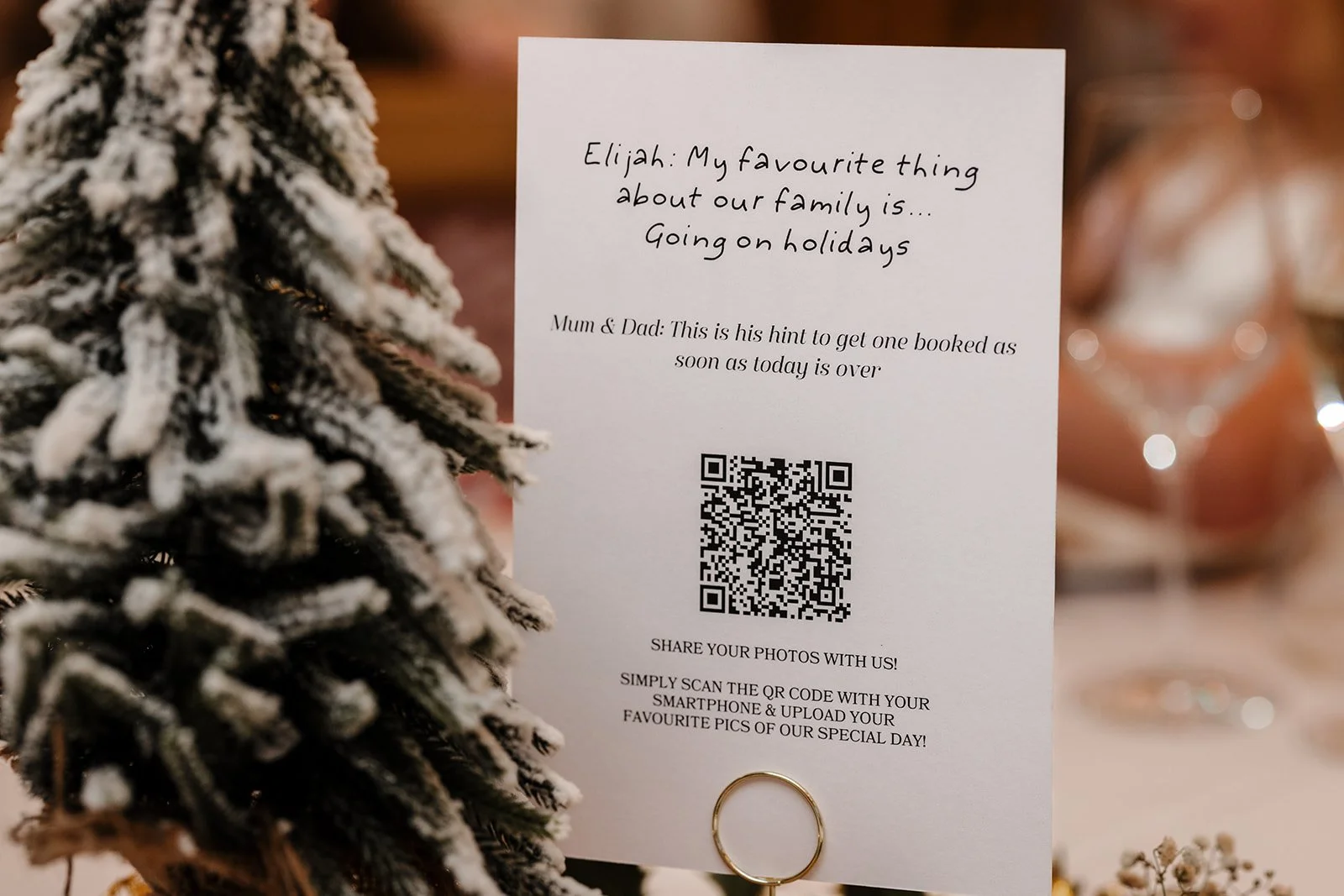Wedding Table Name card mounted on a holder with a QR code, next to a snow-covered Christmas tree, with blurred Christmas decorations in the background.
