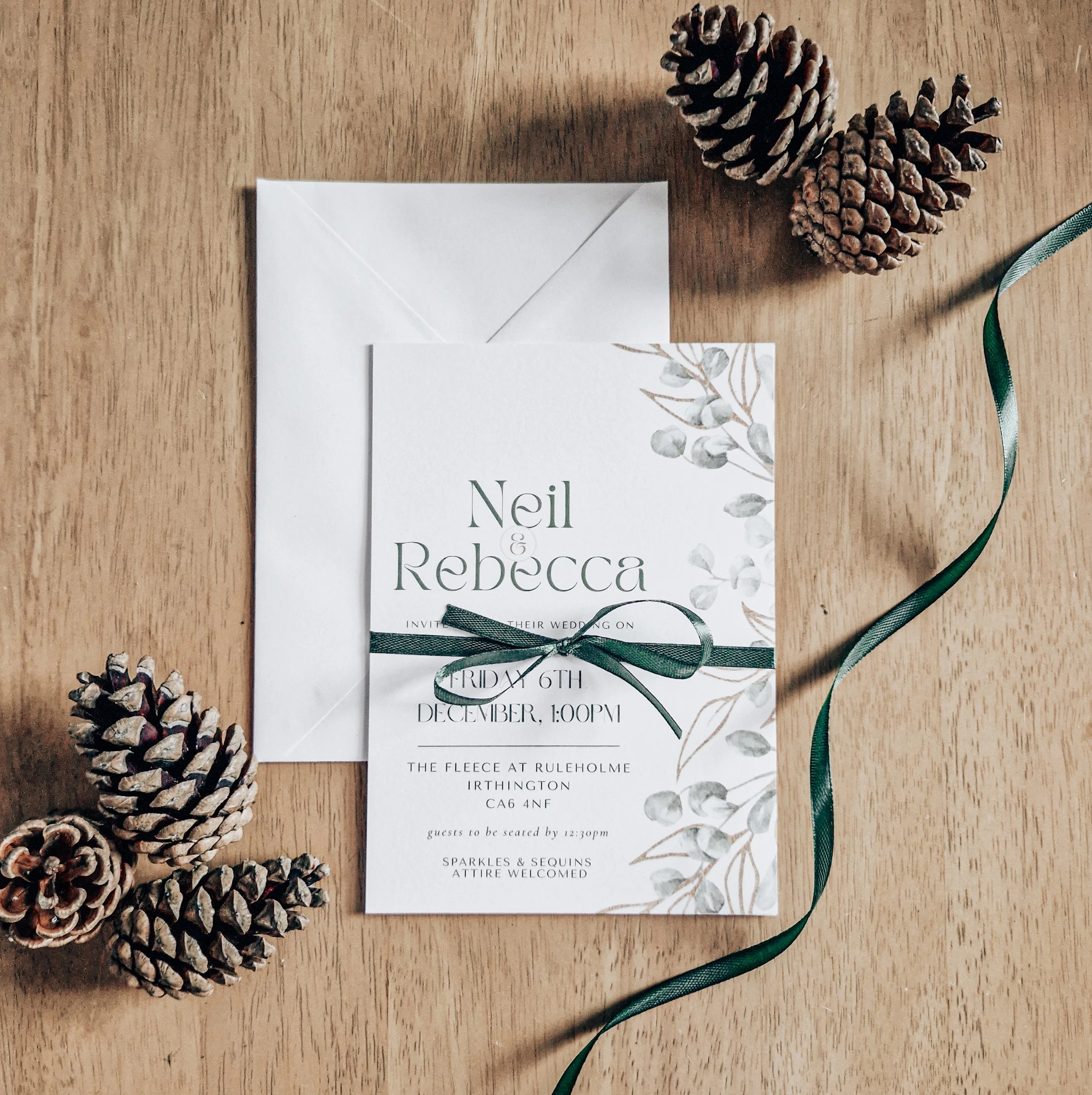Winter Wedding invitation card with green ribbon, surrounded by pinecones, on a wooden surface. The Fleece Ruleholme Carlisle Cumbria