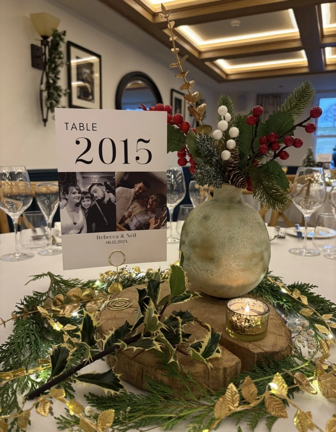A festive table centerpiece featuring a white vase with holiday foliage, berries, and pinecones, surrounded by greenery and a small candle. A table number card displays "TABLE 2015" with photos of Rebecca and Neil, along with their wedding date, 06.1