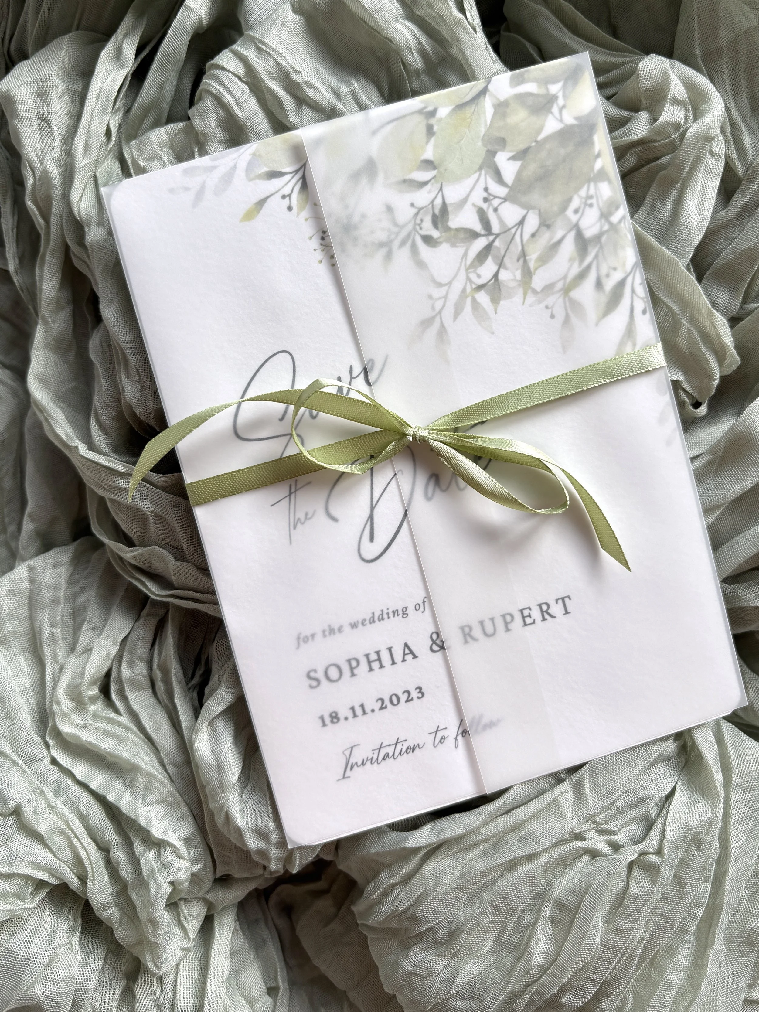 Wedding invitation card with floral design and sage ribbon, placed on soft gray fabric.