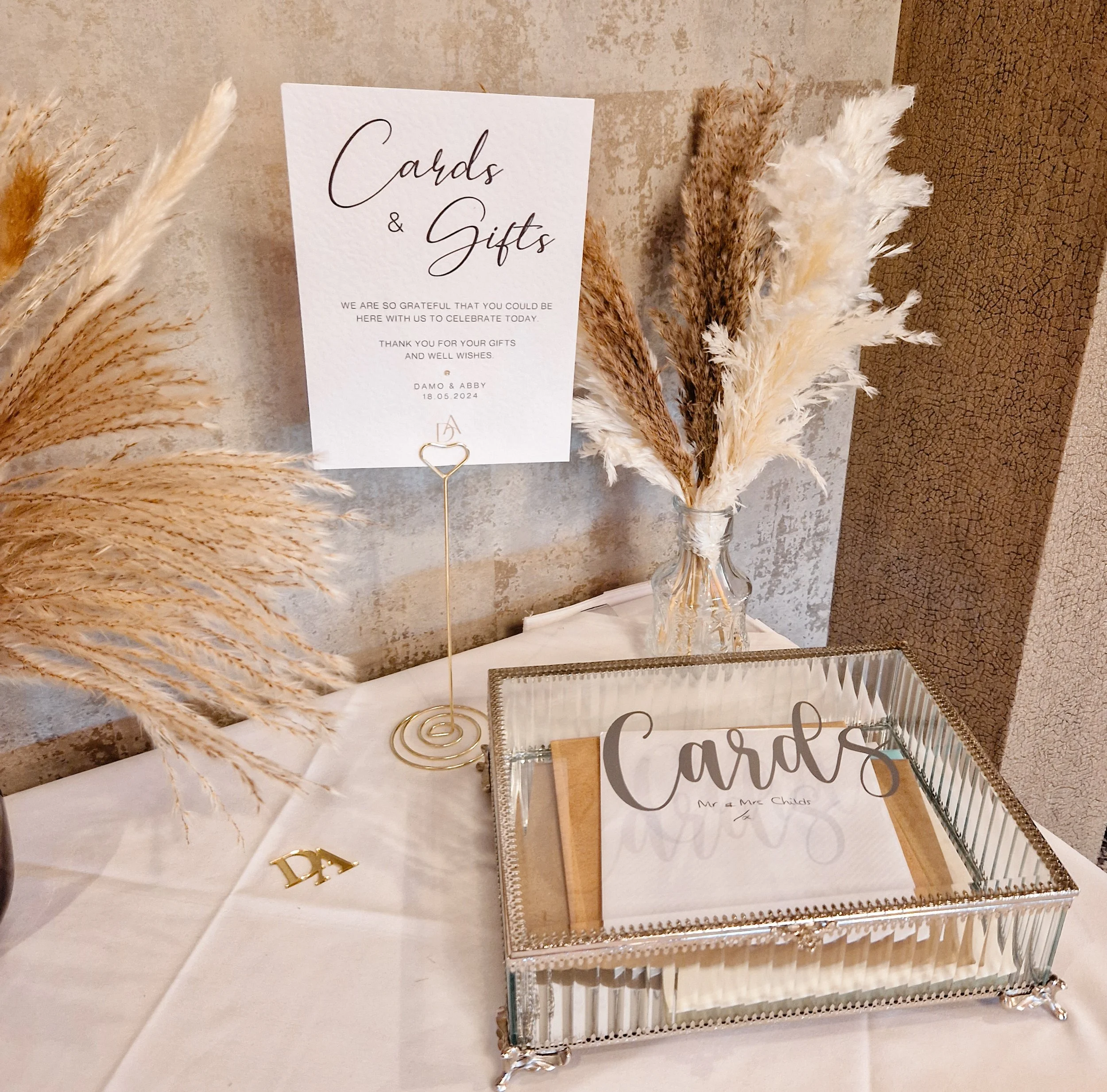 Table with cards and gifts for a celebration, decorated with dried pampas grass in a glass vase, a sign that reads 'Cards & Gifts', a glass box holding cards, and a small gold 'DA' letter