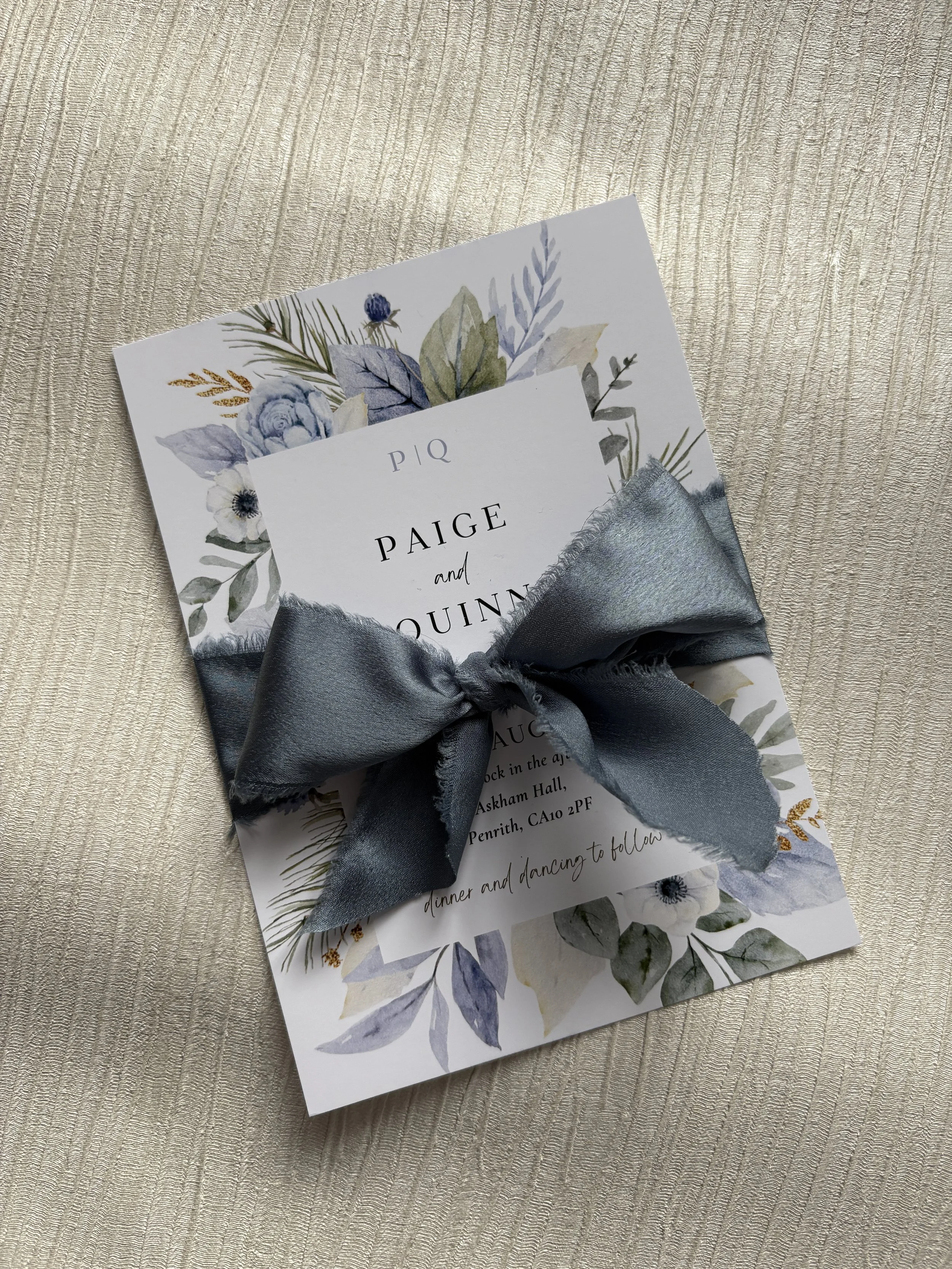 Invitation card with floral design and a dusty blue chiffon ribbon tied in a bow.