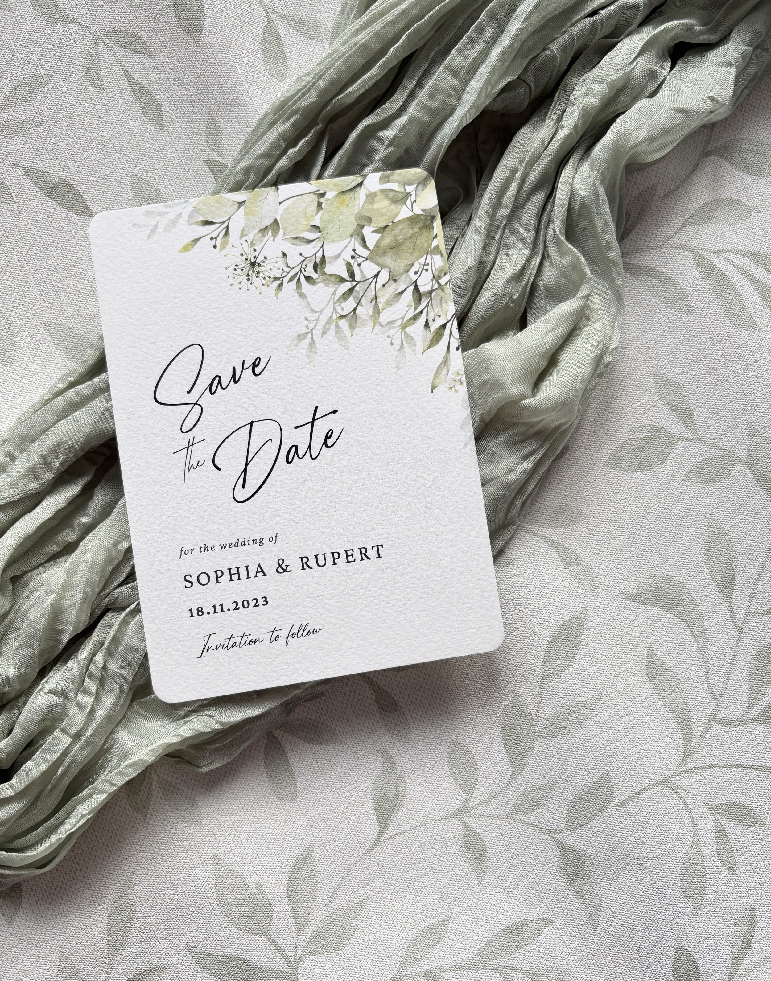 A sage leafy foliage wedding save the date textured card with a green leafy border, placed on a textured surface with a light-colored, leafy pattern cloth underneath.