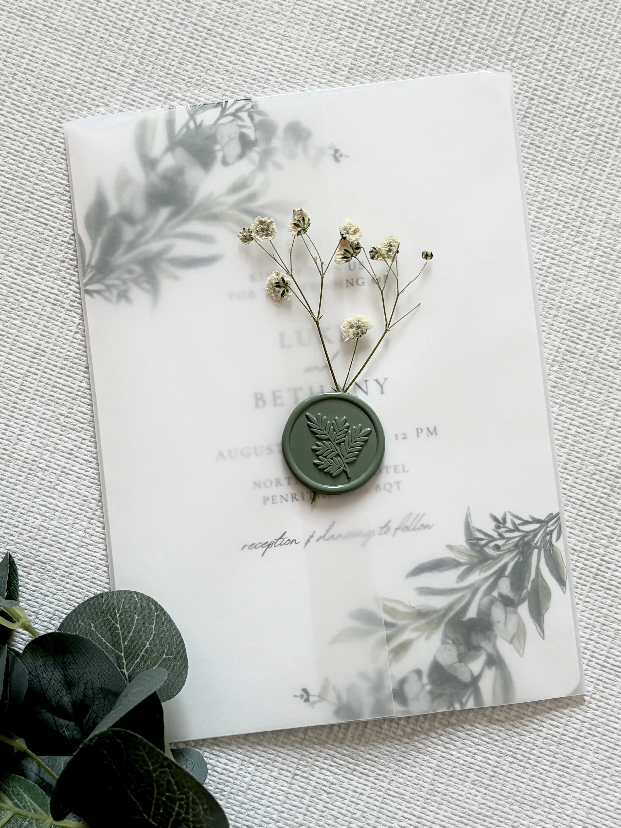 Wedding invitation with floral design and a green wax seal featuring a leaf pattern, adorned with small white flowers and placed on textured fabric.