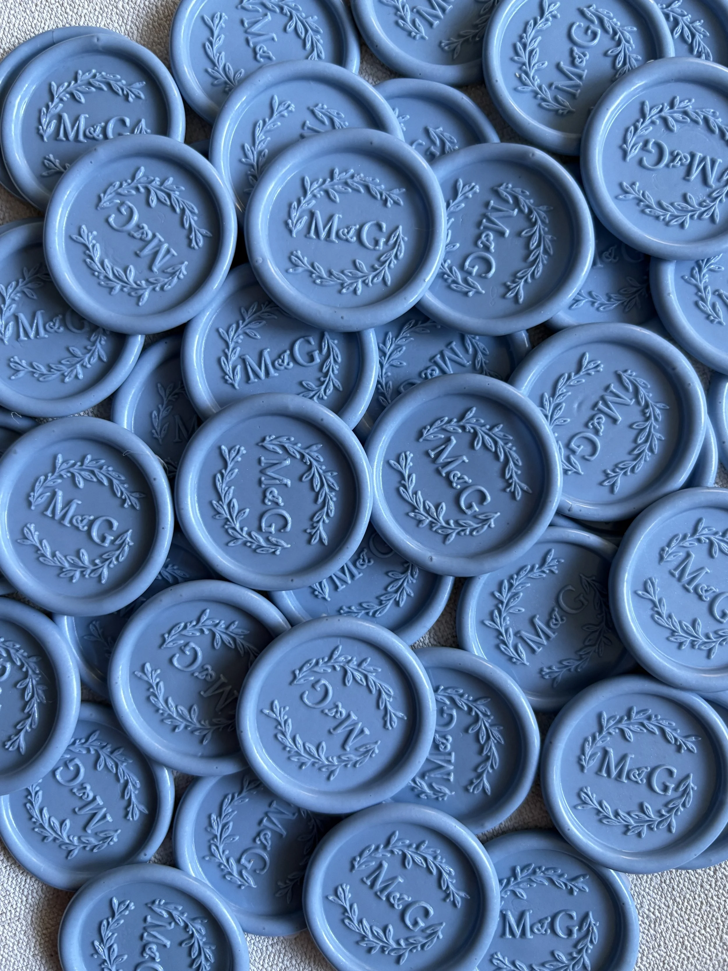 A pile of blue wax seals with custom monogram initials 'M & G' inside a laurel wreath design.