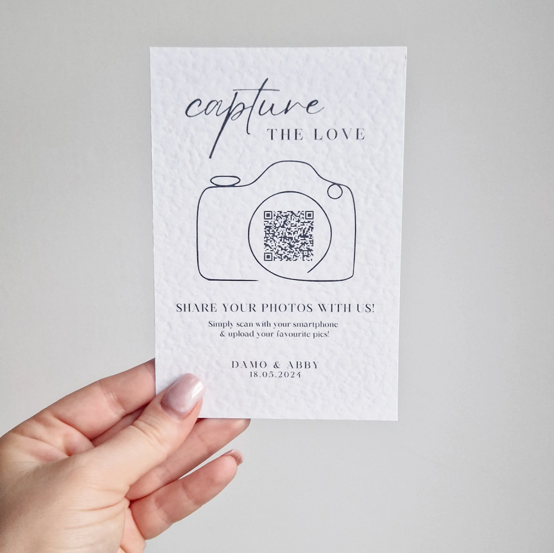 A person holding a white textured paper with a camera outline, QR code, and text for sharing photos, mentioning 'capture the love' and the names Damo & Abby with the date 18.05.2024.