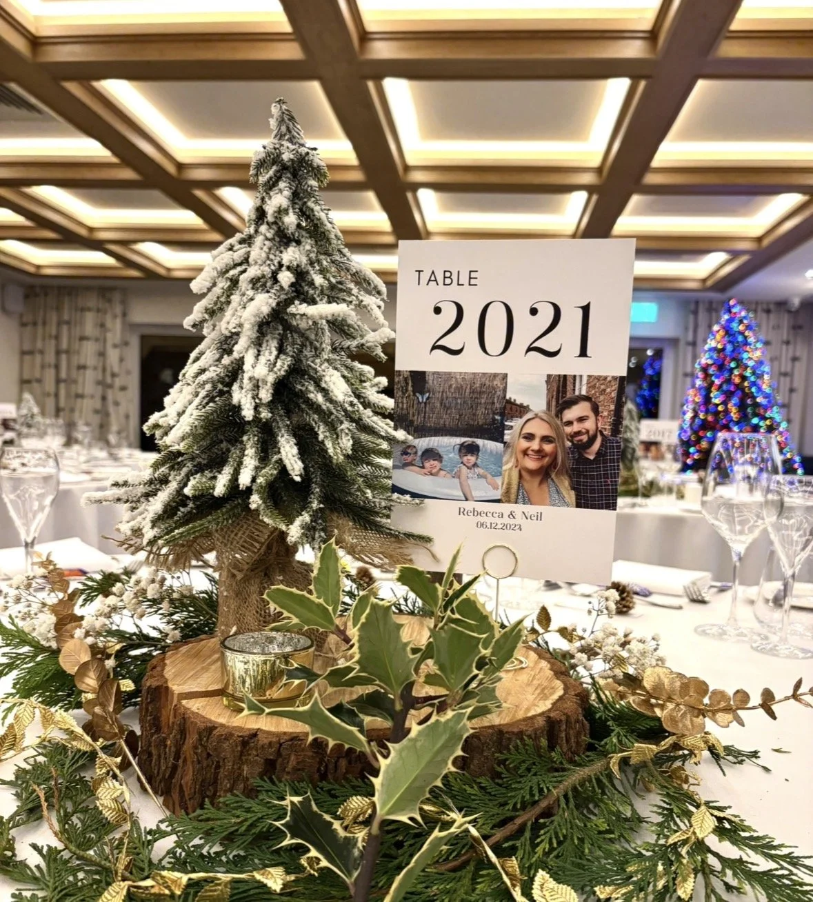 Table centerpiece at a festive event with a small snow-covered Christmas tree, holly, and a framed photo of a couple and two children, with the sign indicating the table is for 2021 and dedicated to Rebecca and Neil, dated December 6, 2023.