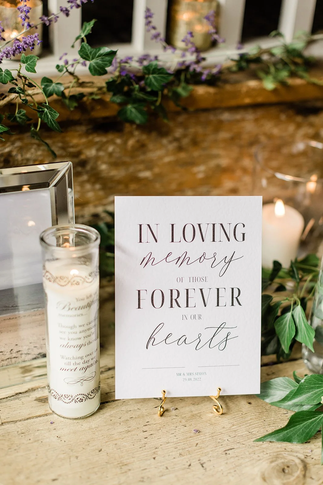 On the Day Wedding Signage reading 'In loving memory of those forever in our hearts,' placed on a wooden table surrounded by candles, greenery, and flowers.