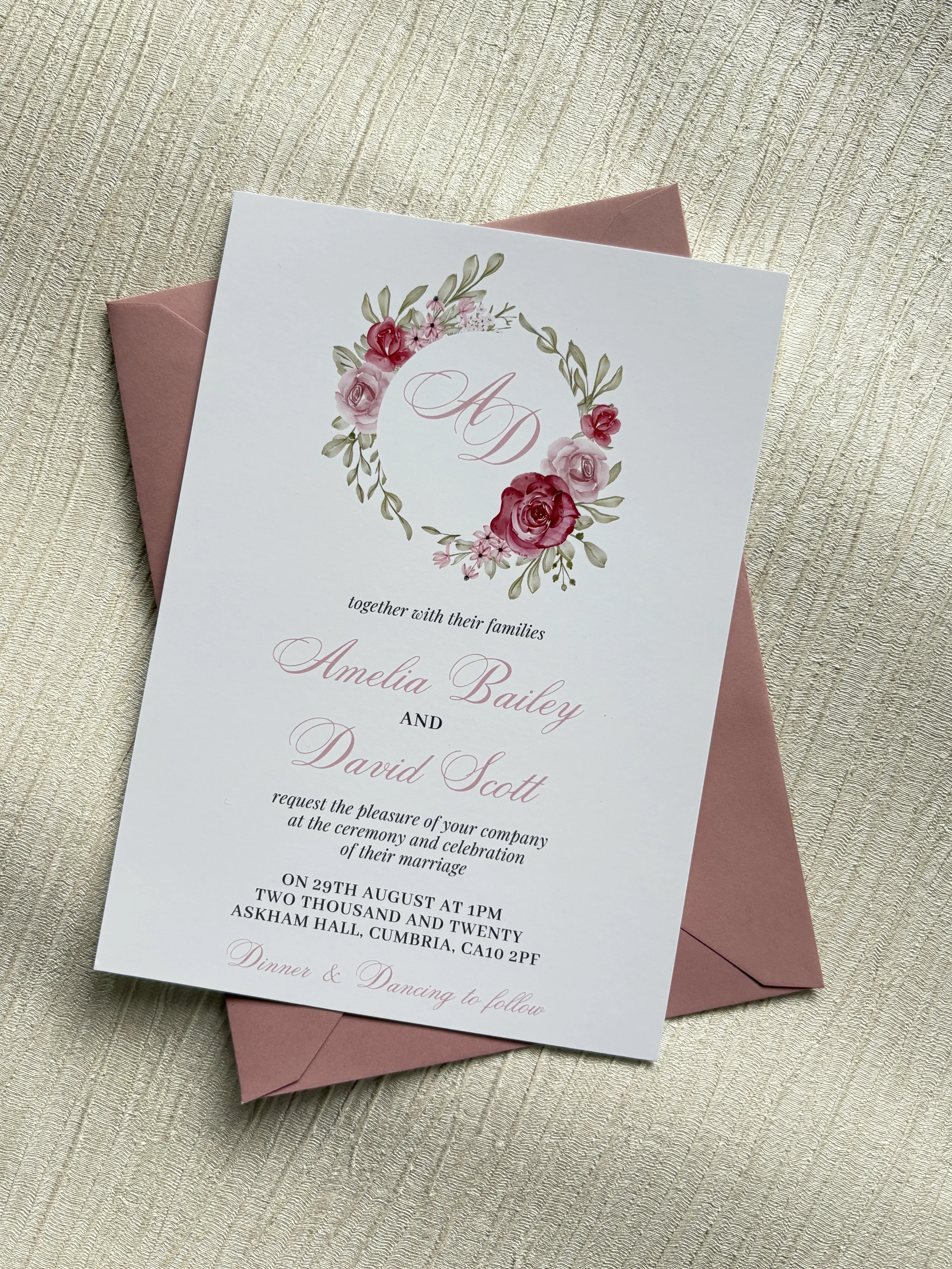 Wedding invitation card with dusky pink floral wreath and pink lettering, placed on dusky pink envelope on a light textured surface.