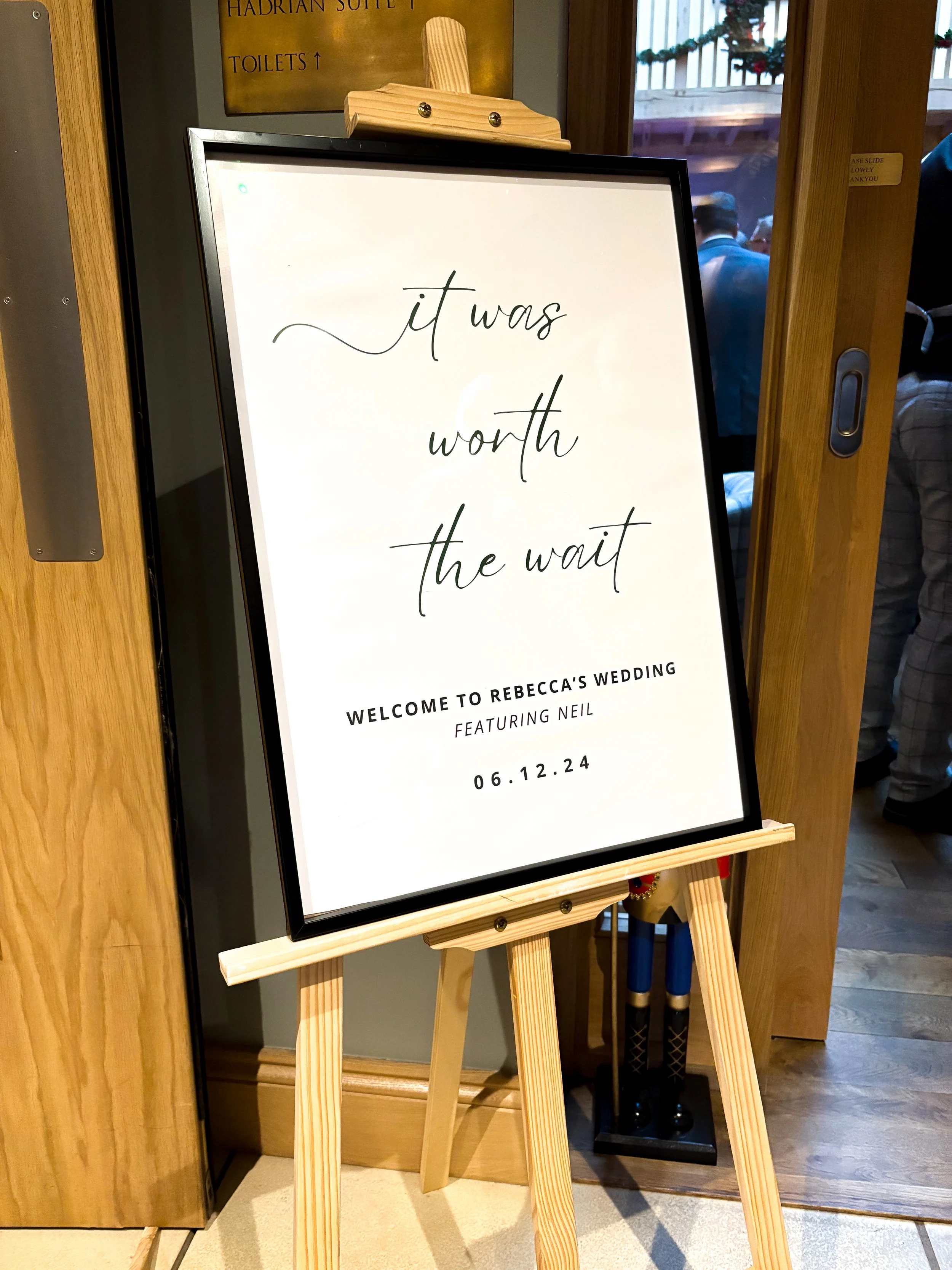 Wedding welcome sign on an easel that reads, 'it was worth the wait. Welcome to Rebecca's wedding featuring Neil, 06.12.24.'