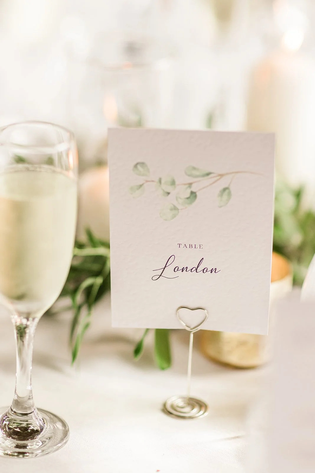 A wedding reception table card with a watercolor green leaf illustration and the text 'Table London'. The card is held upright by a decorative silver heart-shaped holder, with a glass of champagne and blurred greenery in the background.