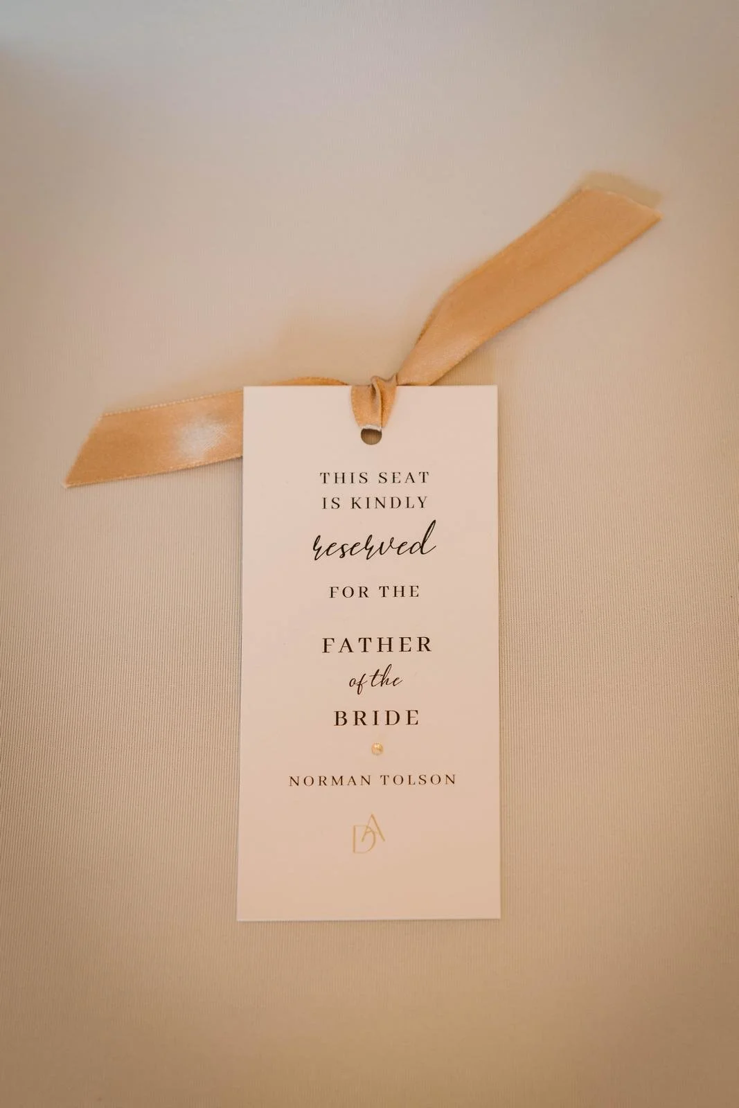 Wedding reserved seat tag with elegant black and gold text attached to an off-white surface with a beige ribbon.