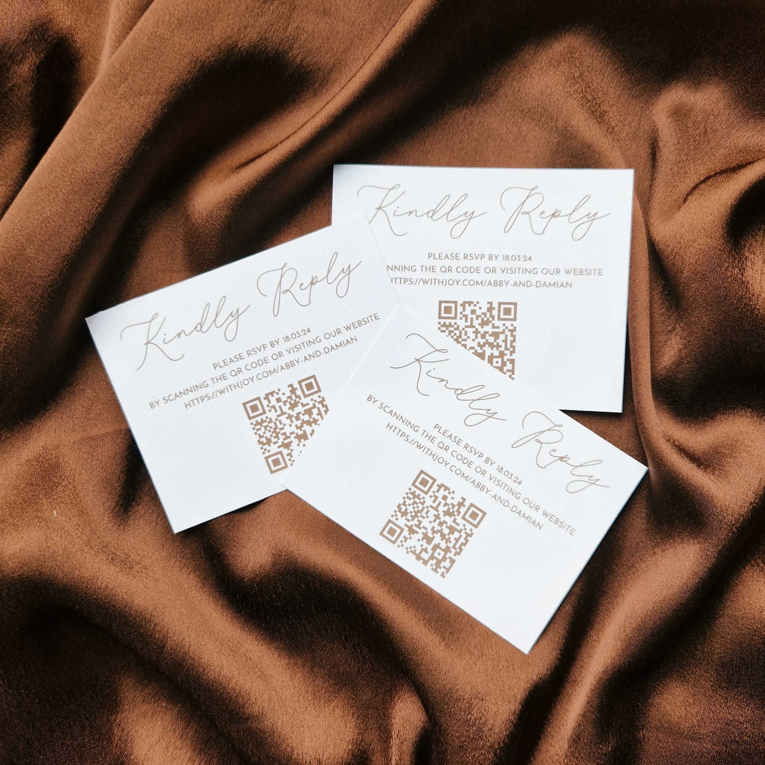 Three white RSVP cards with cursive 'Kindly Reply' text, QR codes, and RSVP instructions, placed on brown satin fabric.
