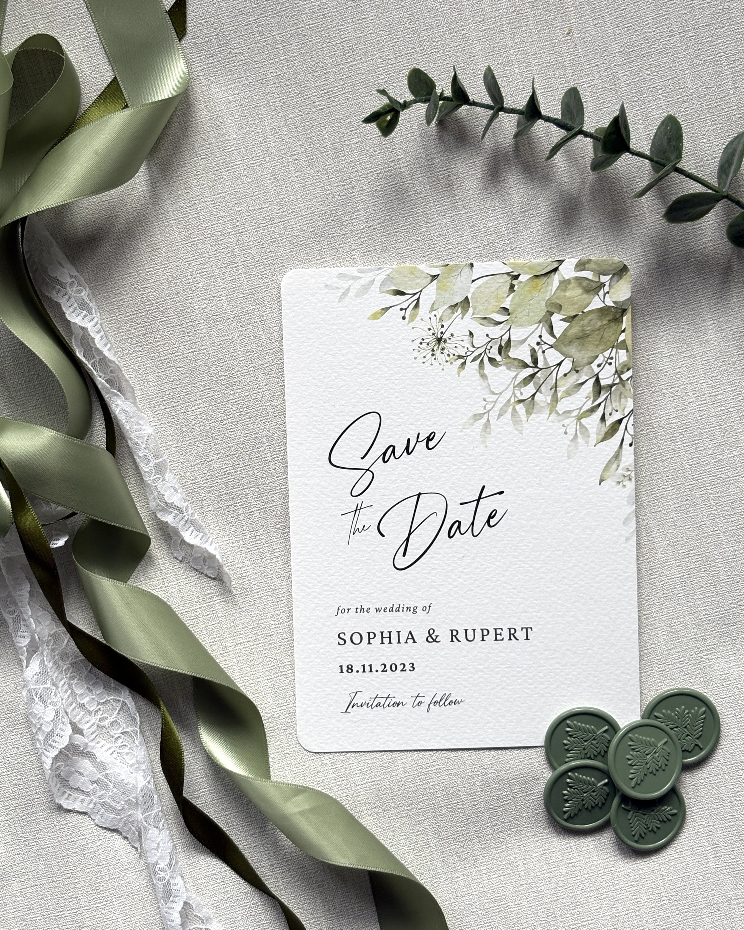 Wedding save the date card on a light-colored fabric surface, decorated with green foliage-themed design, sage olive green sealing wax stamps, green satin and lace ribbons, and a sprig of greenery.