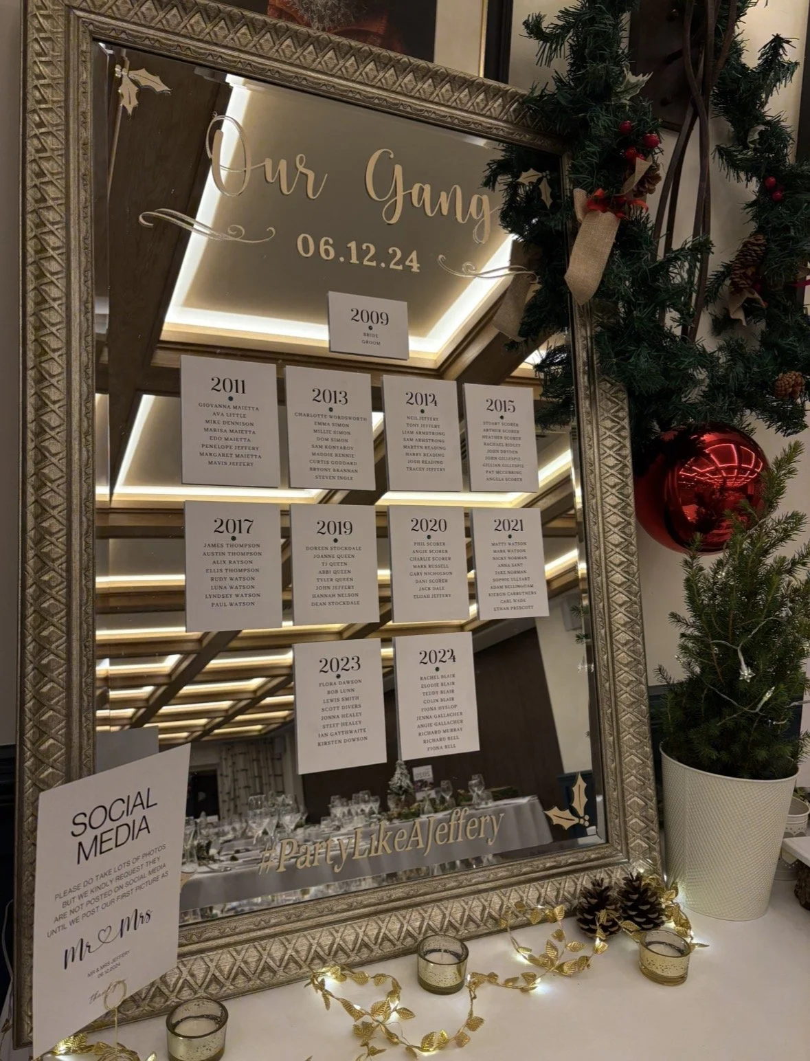 Decorative mirror with a family tree of wedding dates from 2009 to 2024, holiday decorations including pine branches, a red Christmas ornament, pinecones, and small lights, and a social media sign requesting to share photos responsibly.