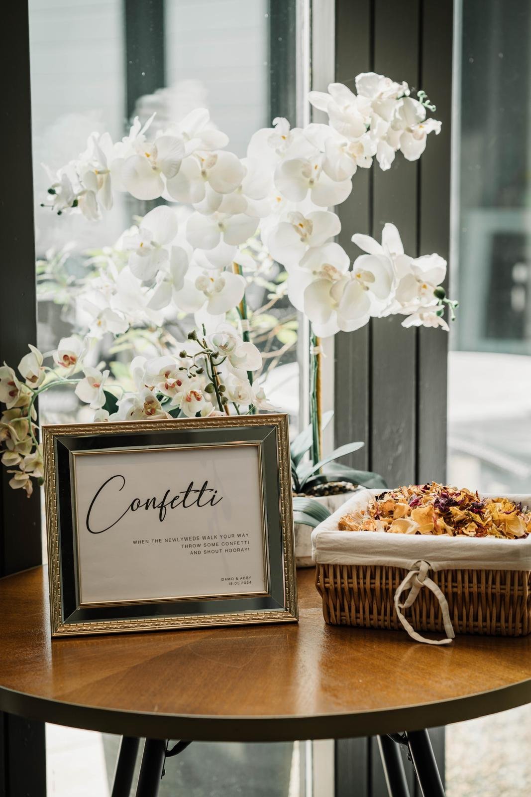 A framed sign that says "Confetti" with a decorative orchid plant and a basket of dried flower petals on a wooden table near a window.