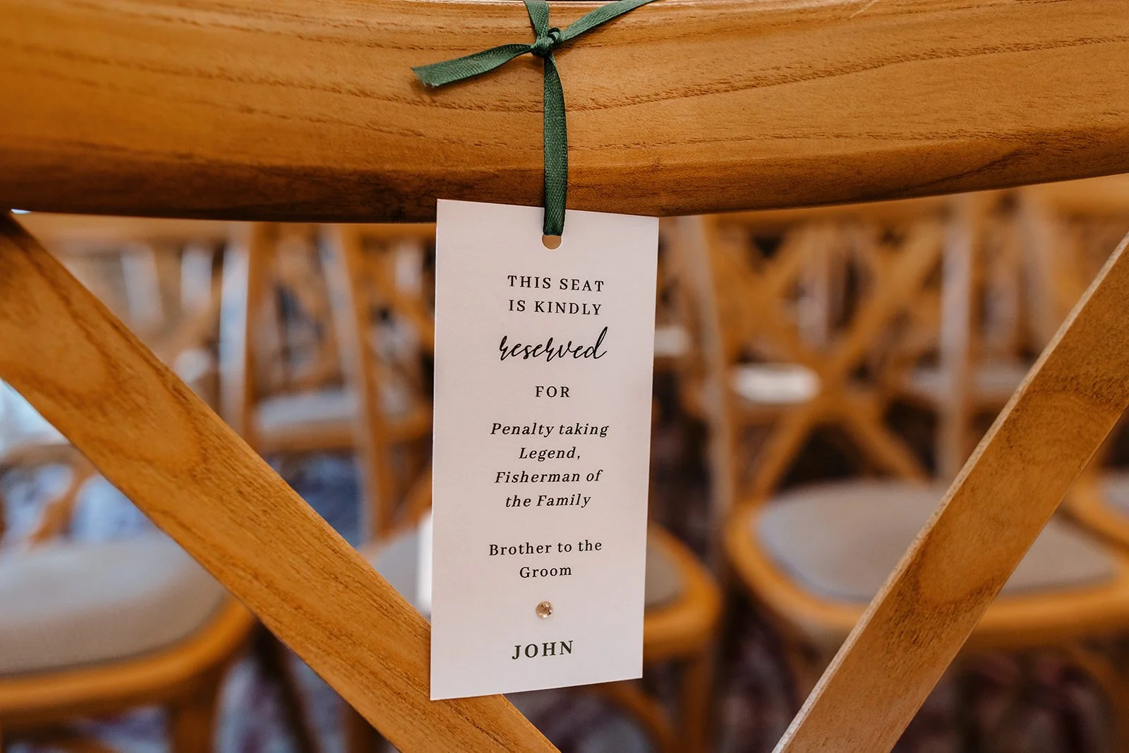 Wedding reserved seat tag hanging from a wooden chair at a wedding. The sign reads, 'This seat is kindly reserved for Penalty taking Legend, Fisherman of the Family, Brother to the Groom, John,' with a small decorative gem at the bottom.