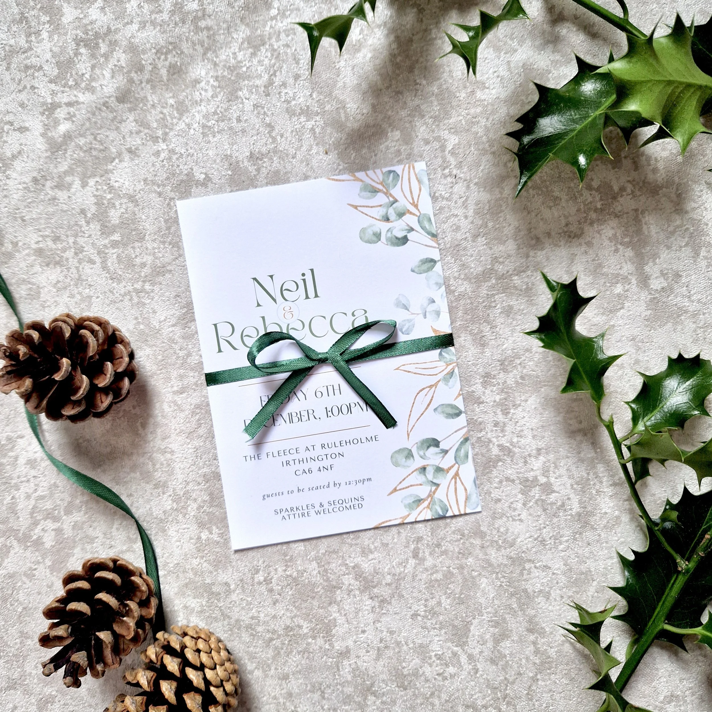 Winter Wedding invitation with green ribbon on a textured surface, surrounded by holly leaves and pinecones. The Fleece Ruleholme Carlisle Cumbria