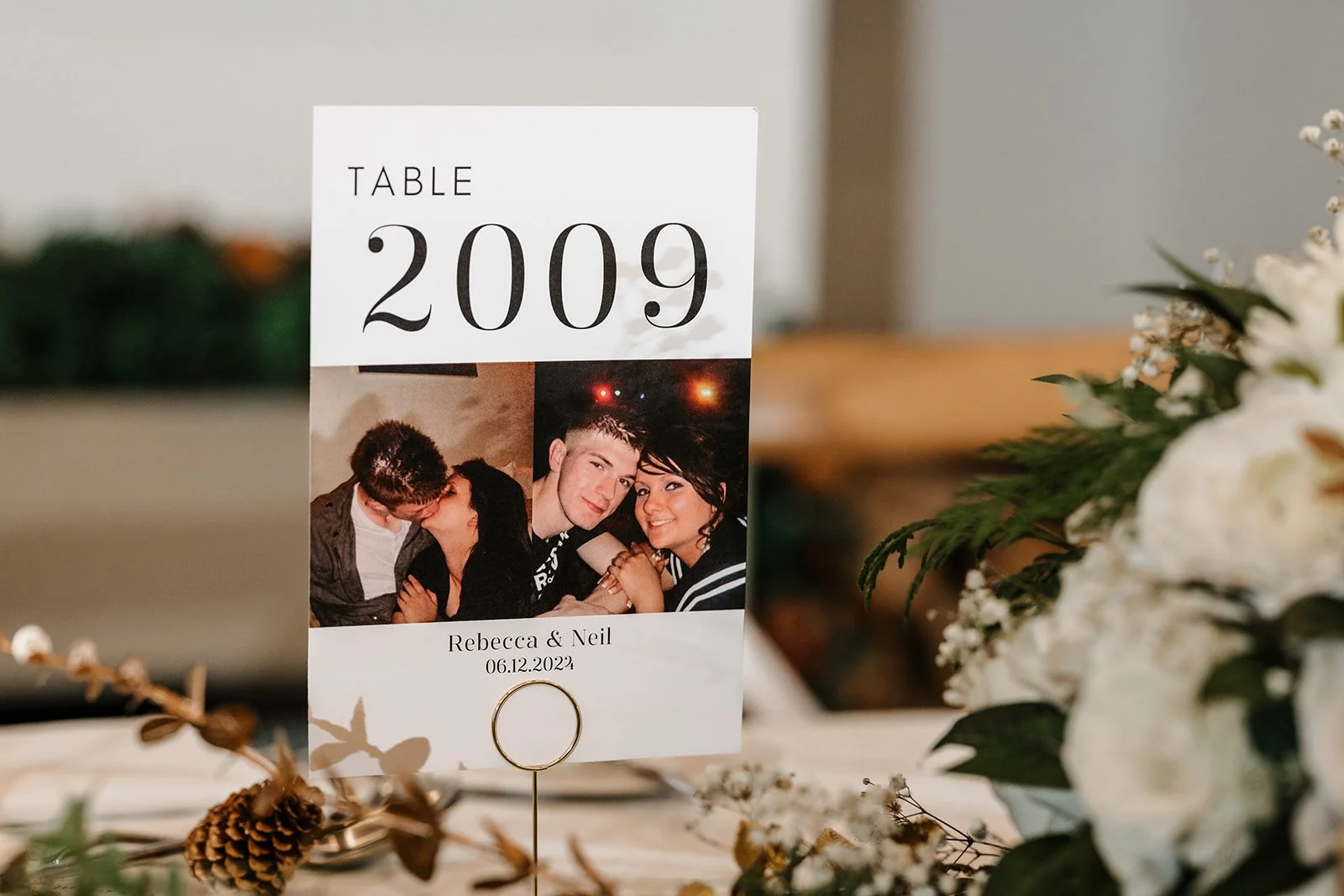 Personalised Table card with the number 2009, a wedding photo of a couple, and text indicating the names Rebecca & Neil and the date 06.12.2024, surrounded by flowers and greenery.