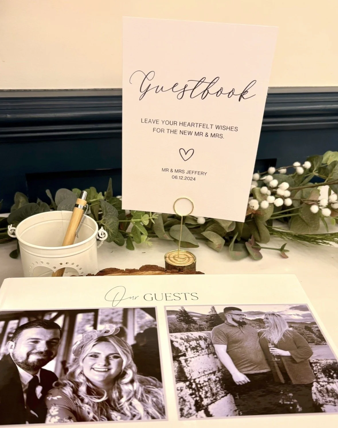 Bespoke Wedding Guestbook sign, guest photos, pen, and decorative greenery with white flowers at a wedding reception.