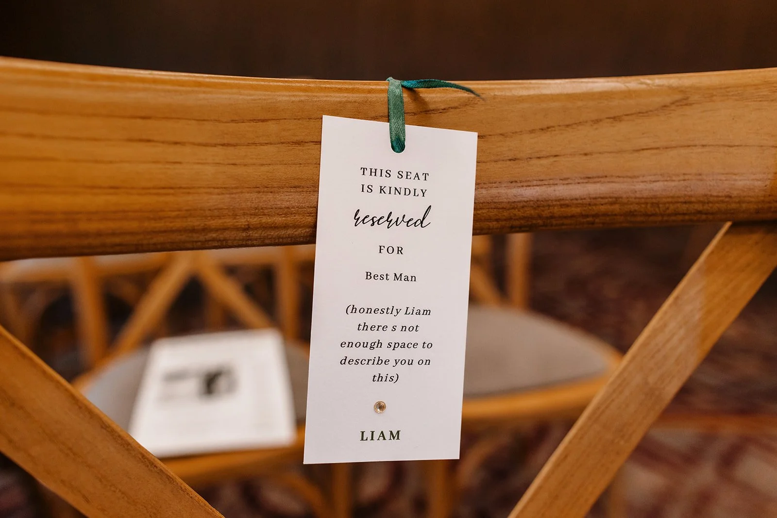 Wooden chair with a wedding reserved seat tag hanging from the backrest. The sign reads: 'This seat is kindly reserved for Best Man. Honestly Liam, there's not enough space to describe you on this. Liam.'