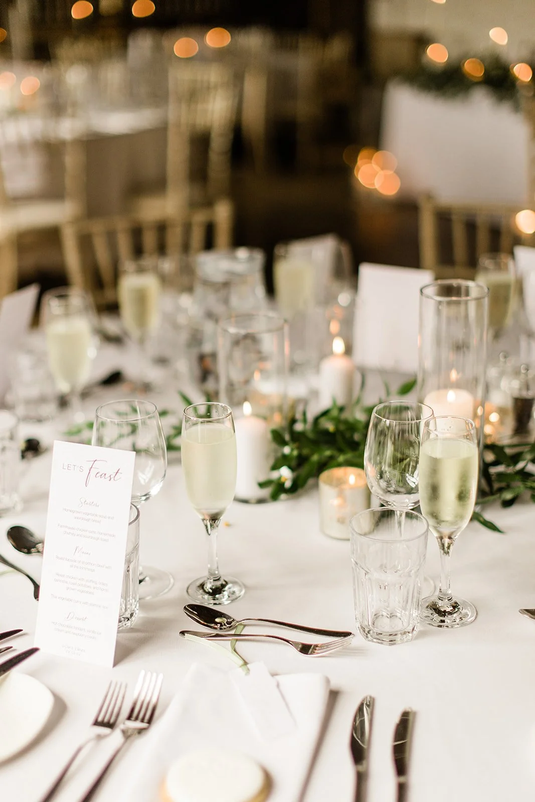 Elegant wedding reception table with white tablecloth, champagne glasses, flickering candles, greenery, and a printed menu, in a warmly lit venue.
