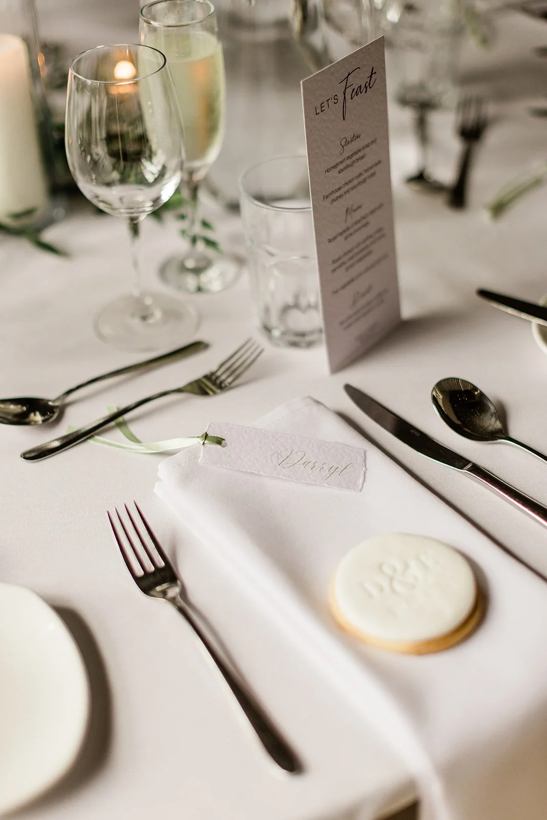 Elegant table setting with silverware, wine glasses, a menu card, a white napkin, and a decorated cookie on a white tablecloth.