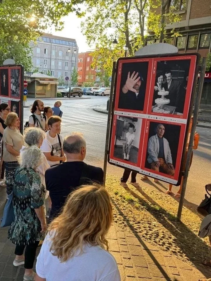 Gruppo di persone che osservano una mostra fotografica all'aperto, con fotografie esposte su pannelli vicino alla strada.