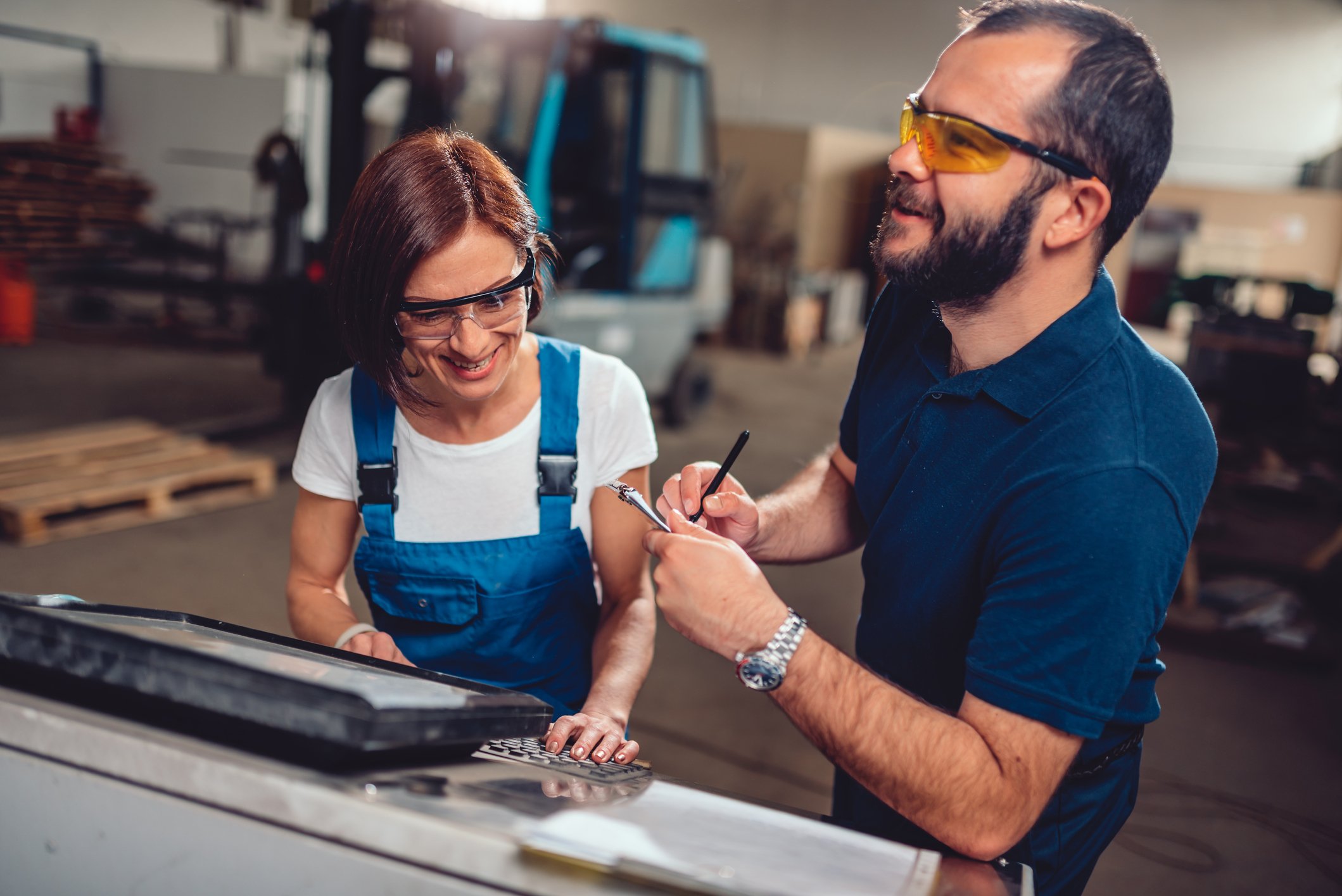 Eine Frau und ein Mann stehen in Arbeitskleidung an einer Maschine in einer Fabrikhalle. Beide tragen Schutzbrillen und lächeln.