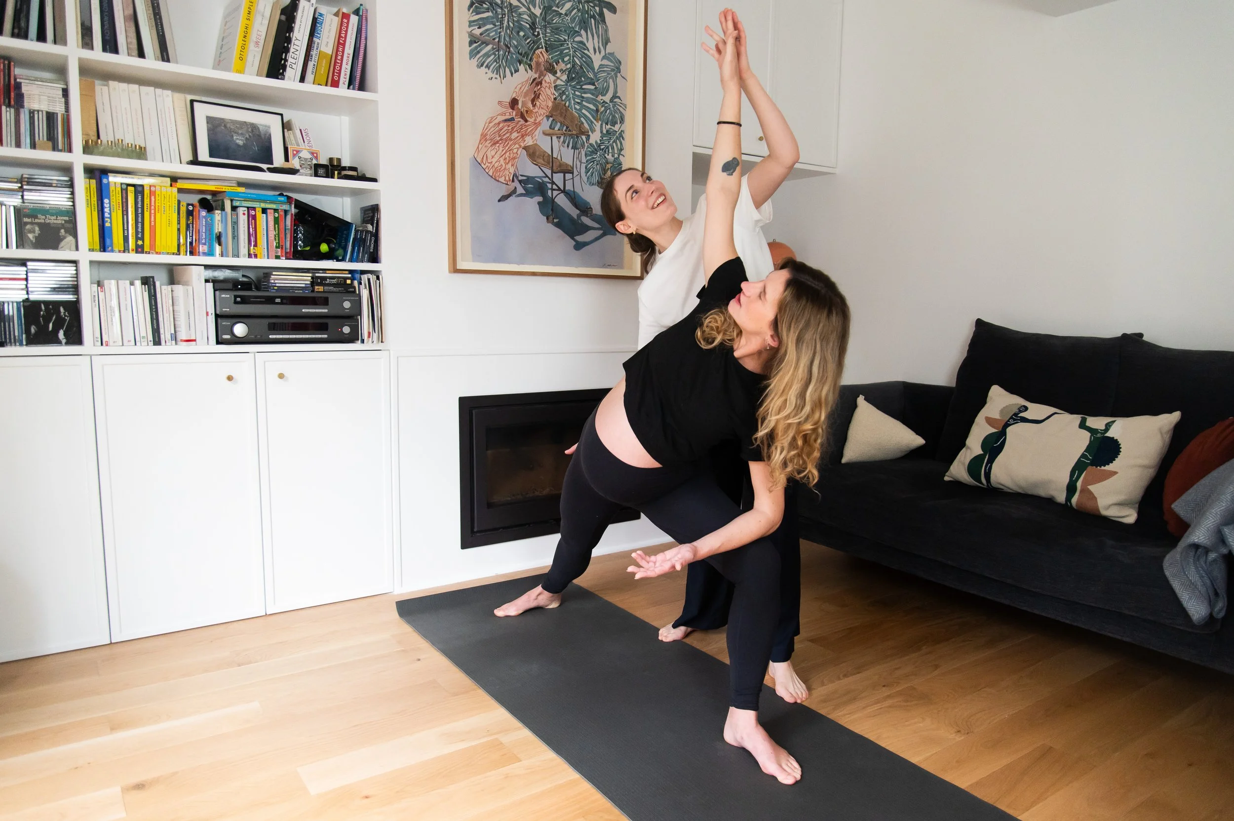 Deux femmes pratiquant le yoga dans un salon, l'une ajuste l'autre dans sa posture, avec une bibliothèque en arrière-plan. Une femme est enceinte, l'autres est sa doula et prof de yoga prénatal à Paris dans le cadre d'un cours privé à domicile.