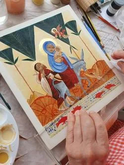 Une personne en train de peindre une icône religieuse dans un livre d'illustrations. La scène représente une figure religieuse, probablement la Vierge Marie avec l'Enfant Jésus, entourée d'arbres et de motifs traditionnels.