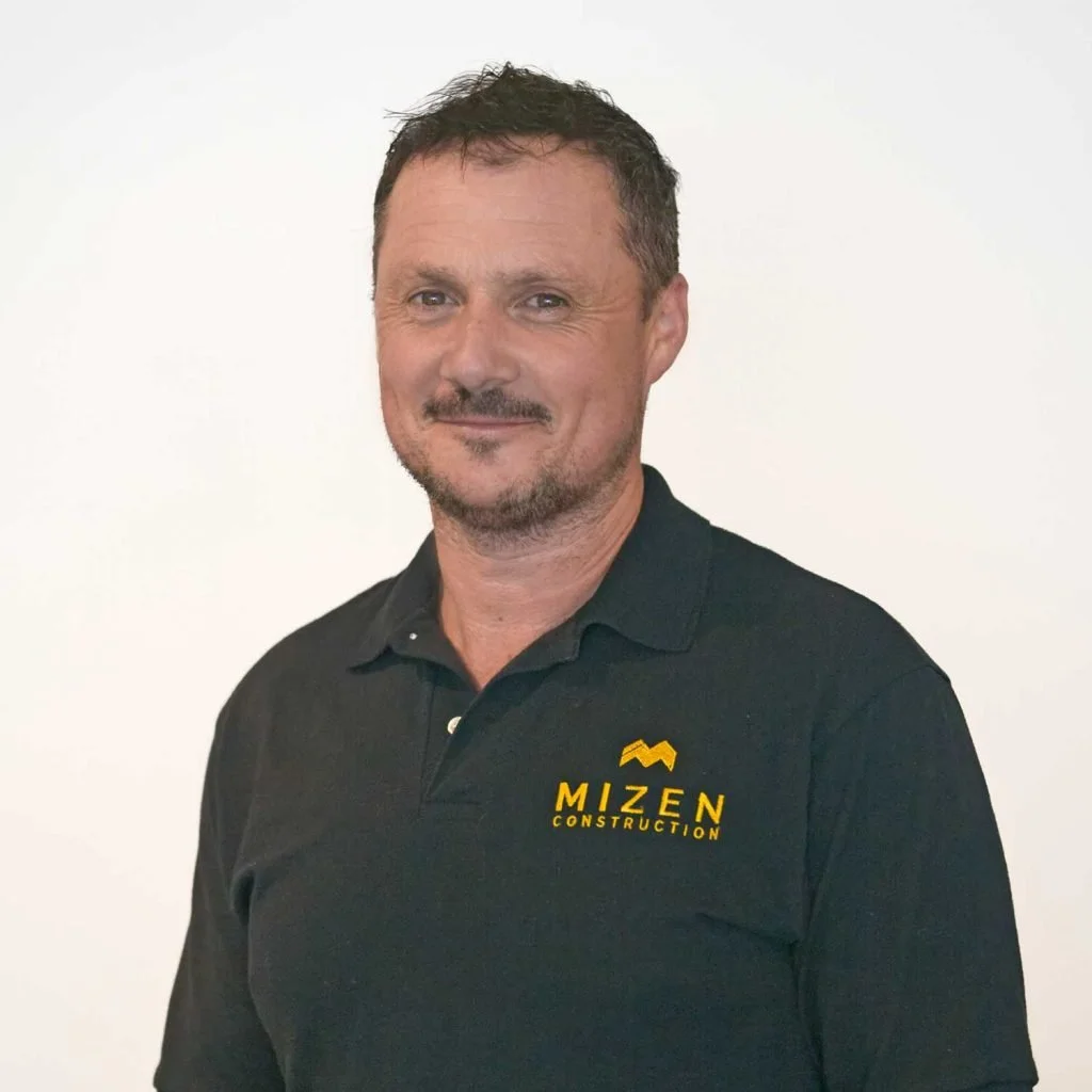 A man with short dark hair, a light beard, wearing a black polo shirt with the yellow and black logo of Mizen Construction, standing against a plain white background.