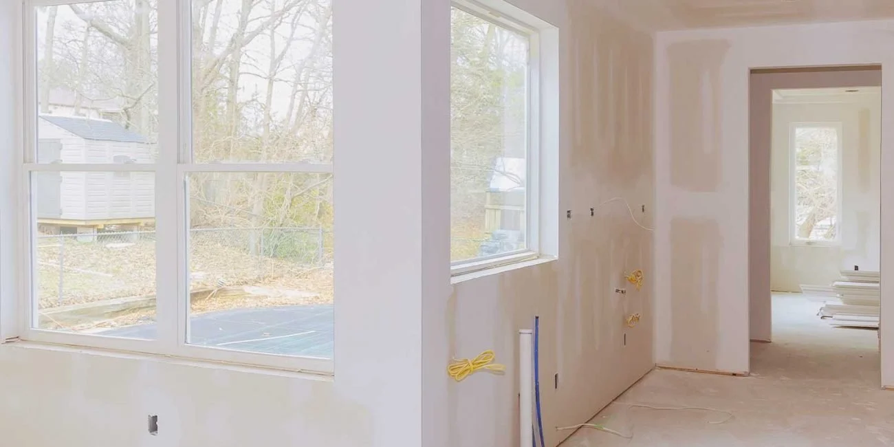Interior of a house under construction, with unfinished drywall and large windows showing an outdoor yard with trees and a shed.