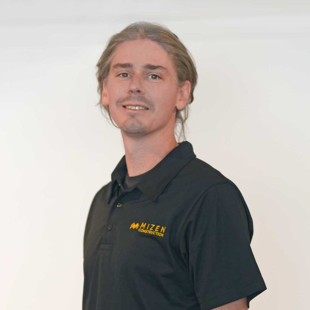 A man with long, wavy gray hair, wearing a black polo shirt with the logo 'Mizen Construction,' standing against a white background.