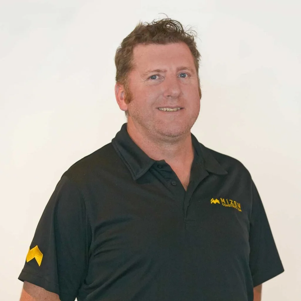 A man with curly light brown hair and blue eyes, smiling, wearing a black polo shirt with a yellow and black logo that reads "MIZEN CONCRETE" on the chest and a yellow emblem on the sleeve, standing against a plain white background.
