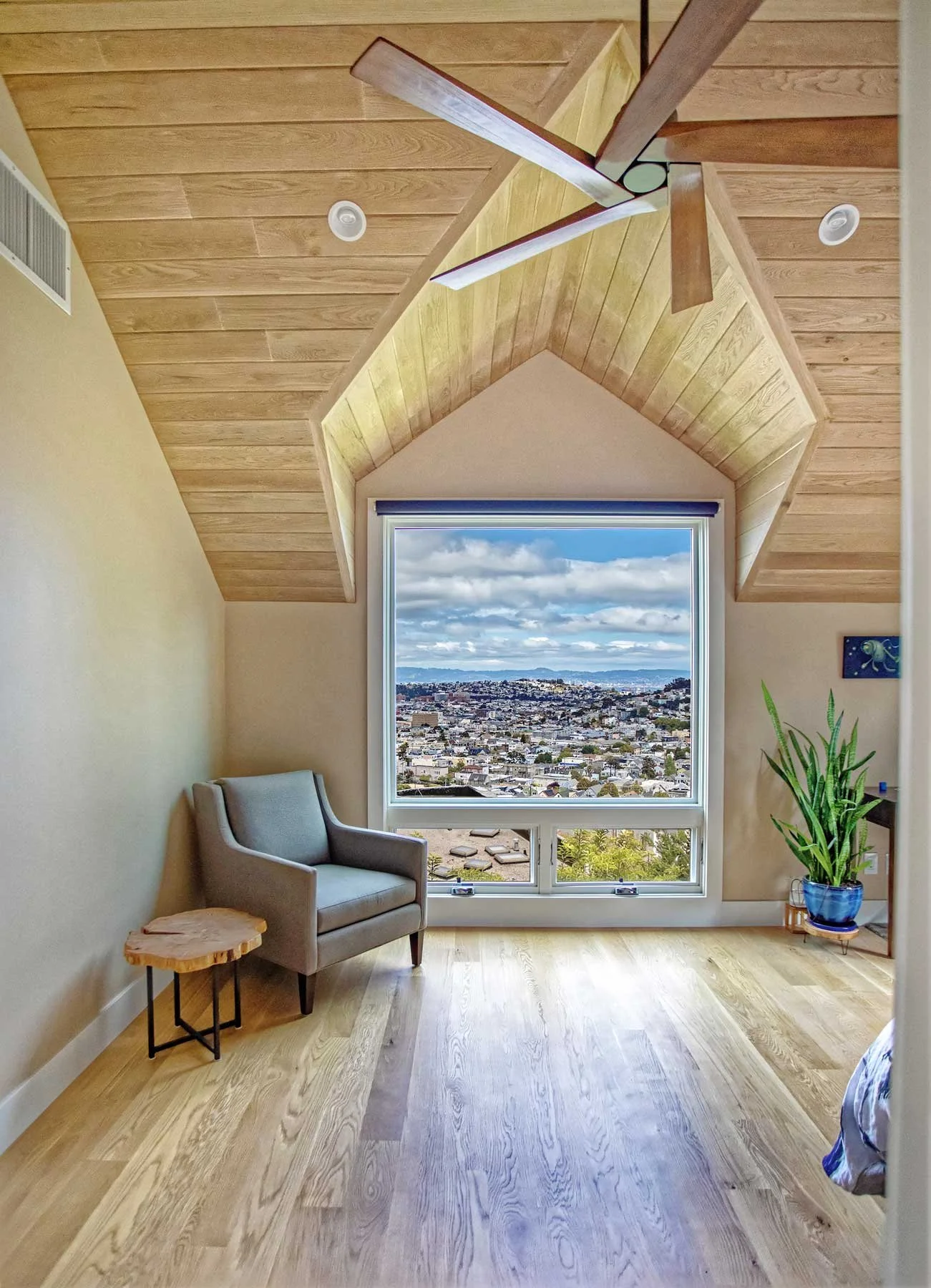 A cozy attic room with a large window overlooking a cityscape, wooden floors, a gray armchair, a small wooden side table, and a potted plant in a blue pot.