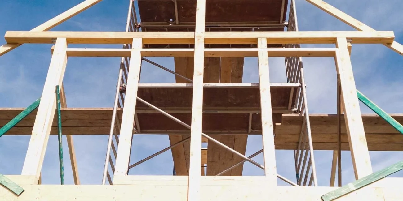 Wooden building framework under construction with scaffolding and a staircase, against a blue sky.