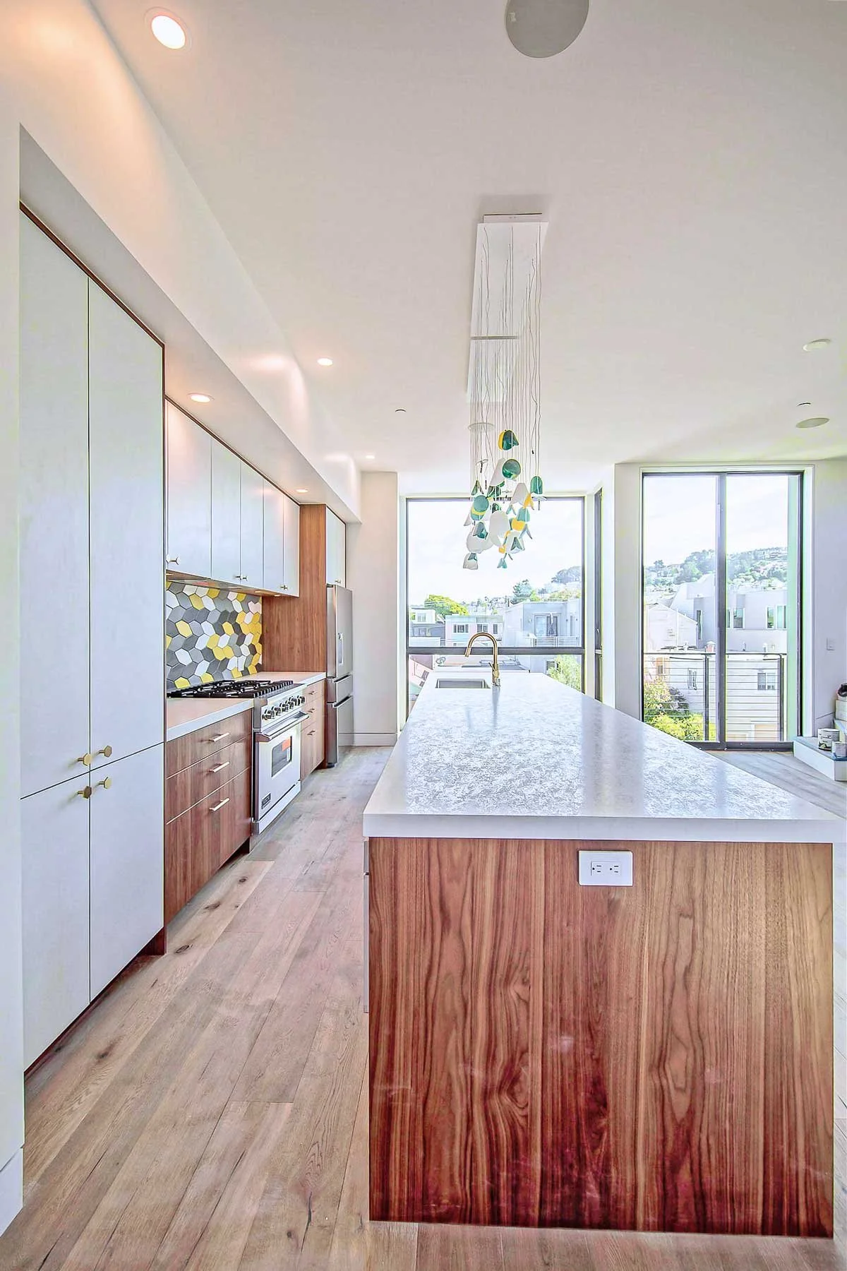 Modern kitchen with wooden cabinets, a large island with a marble countertop, and large windows showing a view of houses and trees outside.
