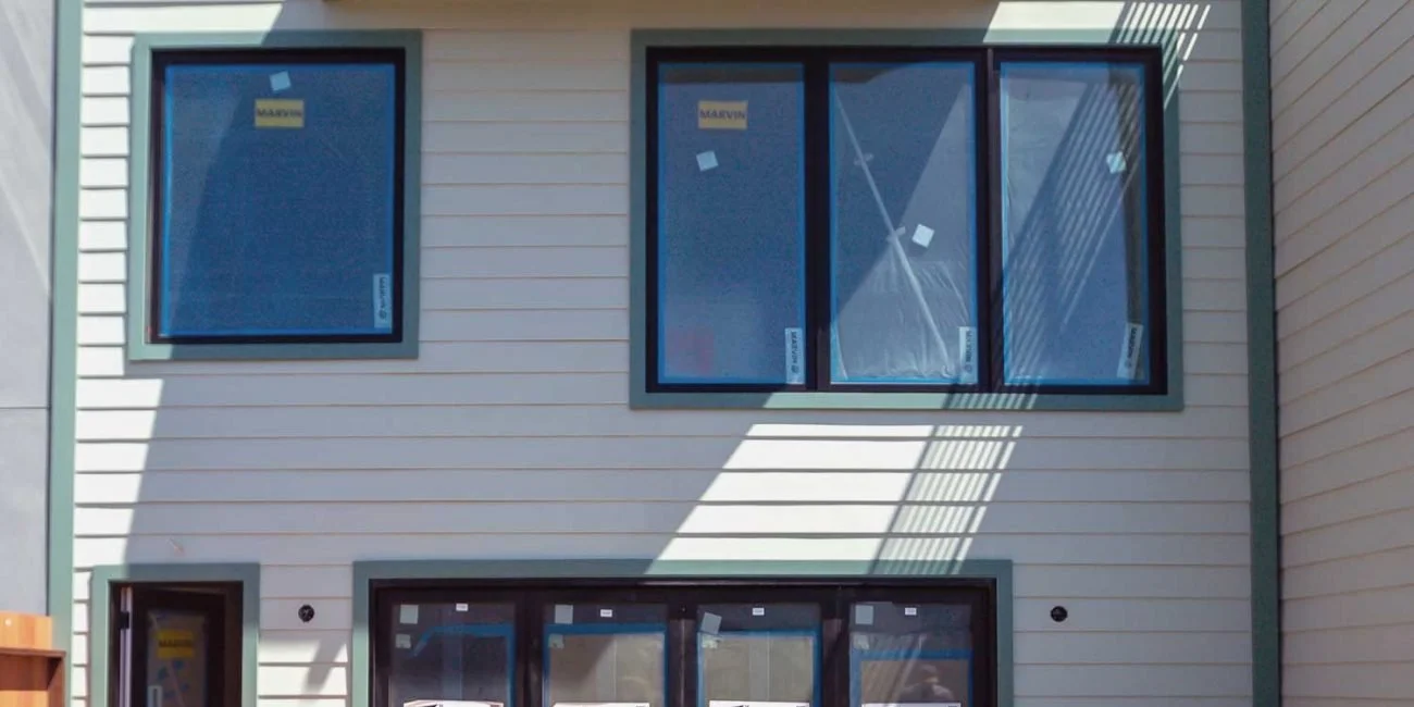 Newly constructed house with beige siding, three large black-framed windows in the upper and lower floors, and a small section of the sky showing reflections on the windows.