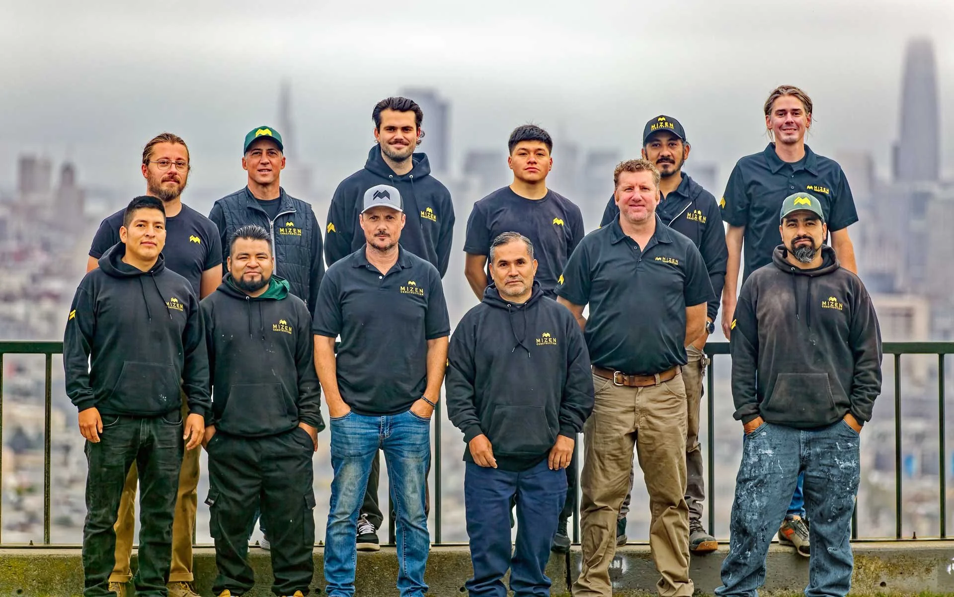 Group of eleven men posing outdoors on a rooftop with city skyline in the background, wearing MIZEN branded clothing and hats.