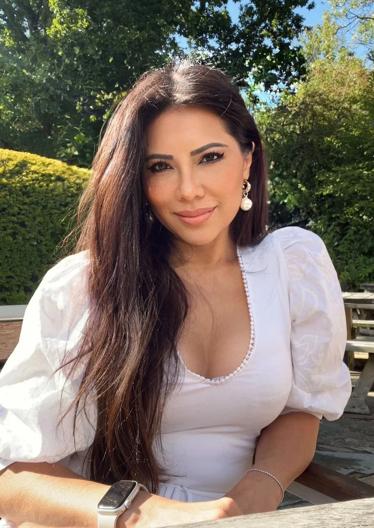 A woman with long dark hair, wearing earrings and a white blouse, sitting outdoors in a park-like setting with greenery and trees in the background.